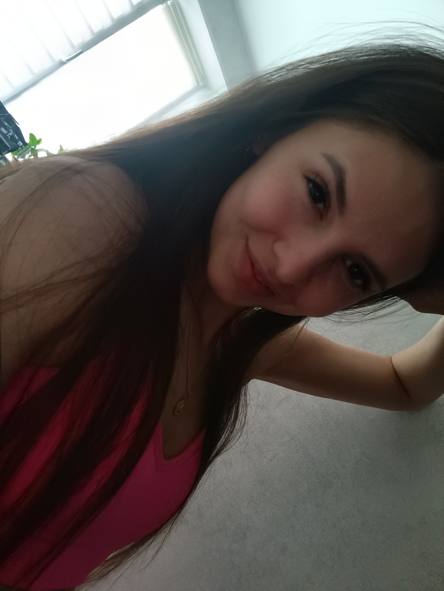 A woman with long, dark hair is leaning against a gray surface. She is wearing a pink tank top and a gold necklace. Light from a window is visible in the upper left corner. The location is Unknown, Unknown.