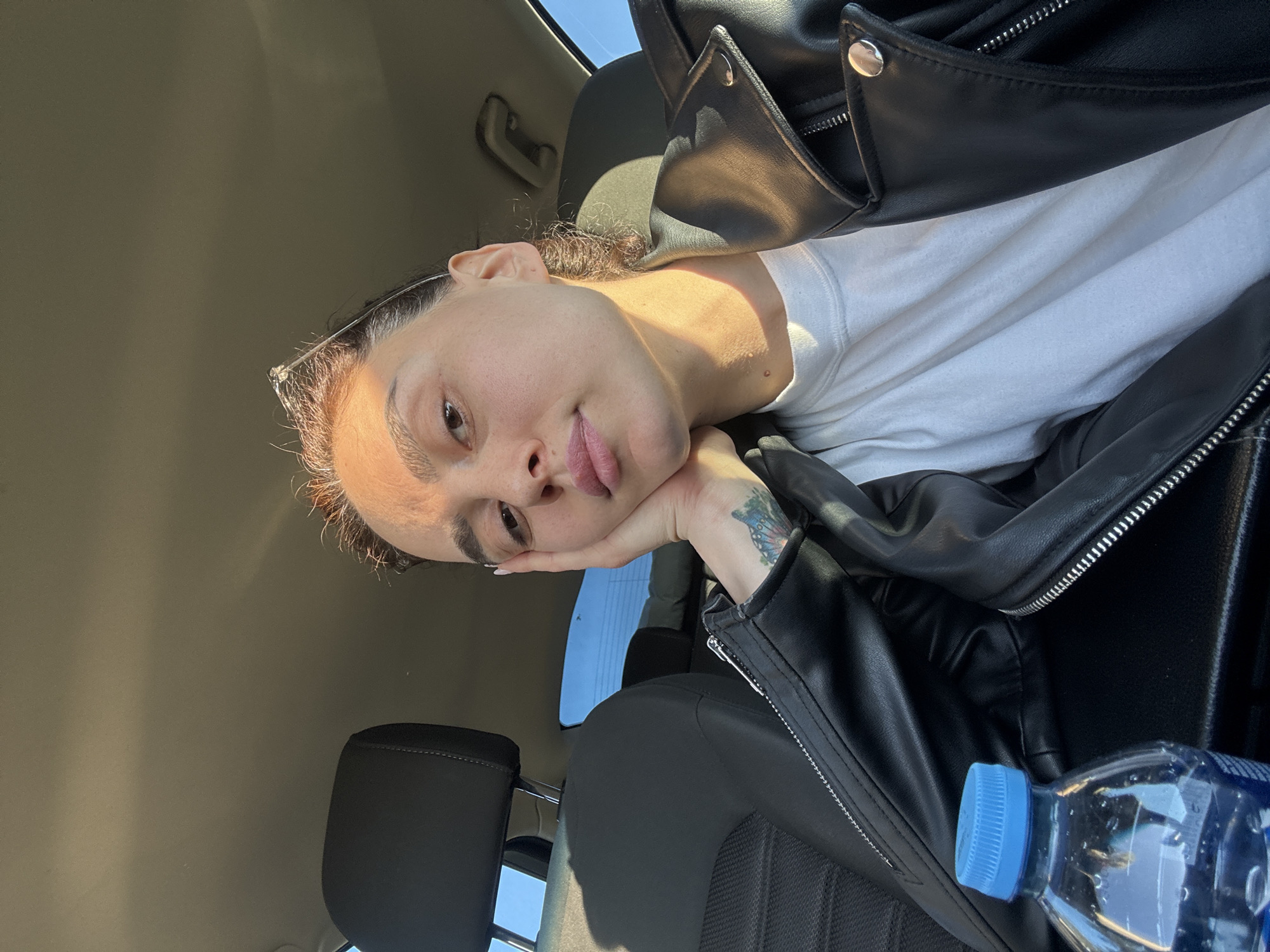 A medium shot captures a person in a car, angled as if taken from an upper vantage point. The individual rests their head on their hand, which bears a tattoo, while gazing towards the camera. They are wearing a white t-shirt underneath a black leather jacket. A bottle of water with a blue cap is visible in the lower right corner. The car's interior is visible, including the seat and headrest. Based on the light and interior, it is impossible to verify the location in the city of Tbilisi, Georgia, without additional contextual information.