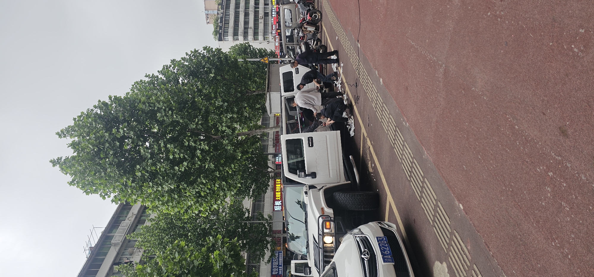 Here is a description of the image.
The image shows a street scene in Yuzhong District, China. A row of white vans are parked along the side of the road. Several men are gathered around one of the vans, appearing to be working on something inside. A white car with the license plate "A-62263" is parked in front of the vans. A sign with Chinese characters is visible in the background, including the characters “太阳膜” (solar film). The road is red and has a textured tactile paving border. Trees are visible on the left side of the image. In the upper background there is a building.