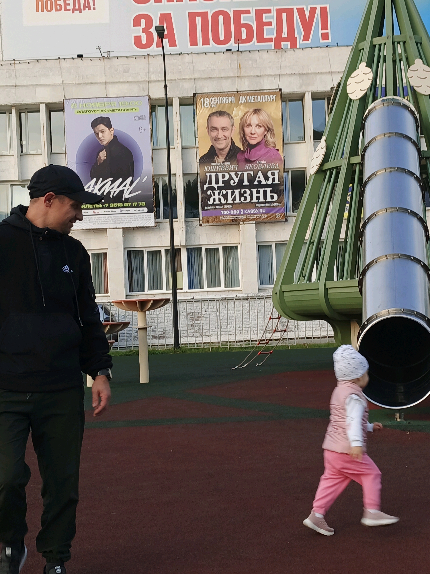 The image captures a segment of a playground with a large, green tubular slide structure dominating the right side. In the background, a building with multiple windows is visible, adorned with large banners. One banner prominently displays the red text "ЗА ПОБЕДУ!" (For Victory!). Another banner features a portrait of a young man with the name "AKMAAL" below, and the text "18 НОЯБРЯ 19:00 ЗЛАТОУСТ ДК «МЕТАЛЛУРГ»" (November 18, 7:00 PM Zlatoust DK "Metallurg"). This indicates the location is Zlatoust, Russia, at the Palace of Culture "Metallurg". A third banner shows portraits of a man and a woman with the text "ДРУГАЯ ЖИЗНЬ" (Another Life). In the foreground, a man wearing a black hoodie and a black baseball cap is seen from the side, looking down and to his right, with his left arm slightly bent. To the right of the man, a young child in a pink vest and pink pants walks away from the camera. The playground surface is a dark reddish-brown.