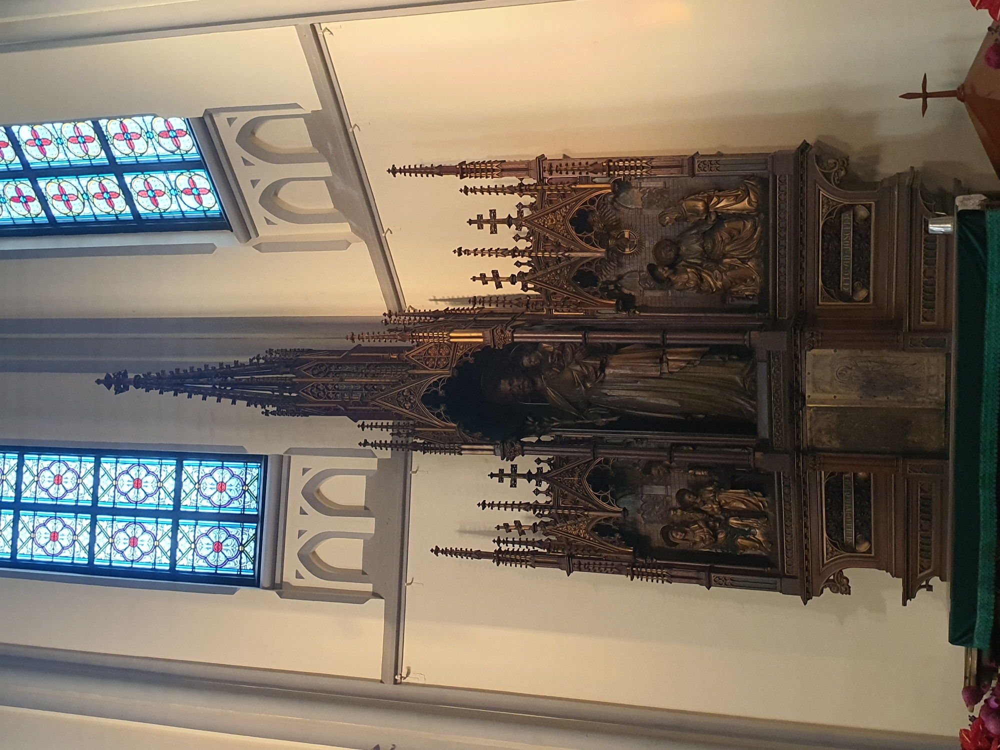 This is a medium shot, taken from a slightly low angle, of a grand altar in what appears to be a church. The altar is made of dark wood with intricate carvings and ornate Gothic architectural elements, including spires and tracery. In the center, a large sculpture depicts a robed figure holding a child, likely a religious scene. Flanking this central piece are smaller relief sculptures depicting groups of figures. Above the altar, two tall stained-glass windows with geometric patterns in blue, red, and white are visible against a cream-colored wall. The overall impression is one of solemnity and historical grandeur. The image is likely located in the Church of Our Lady of the Assumption in Central Jakarta, Indonesia, a place of worship known for its historical significance and ornate religious artistry.