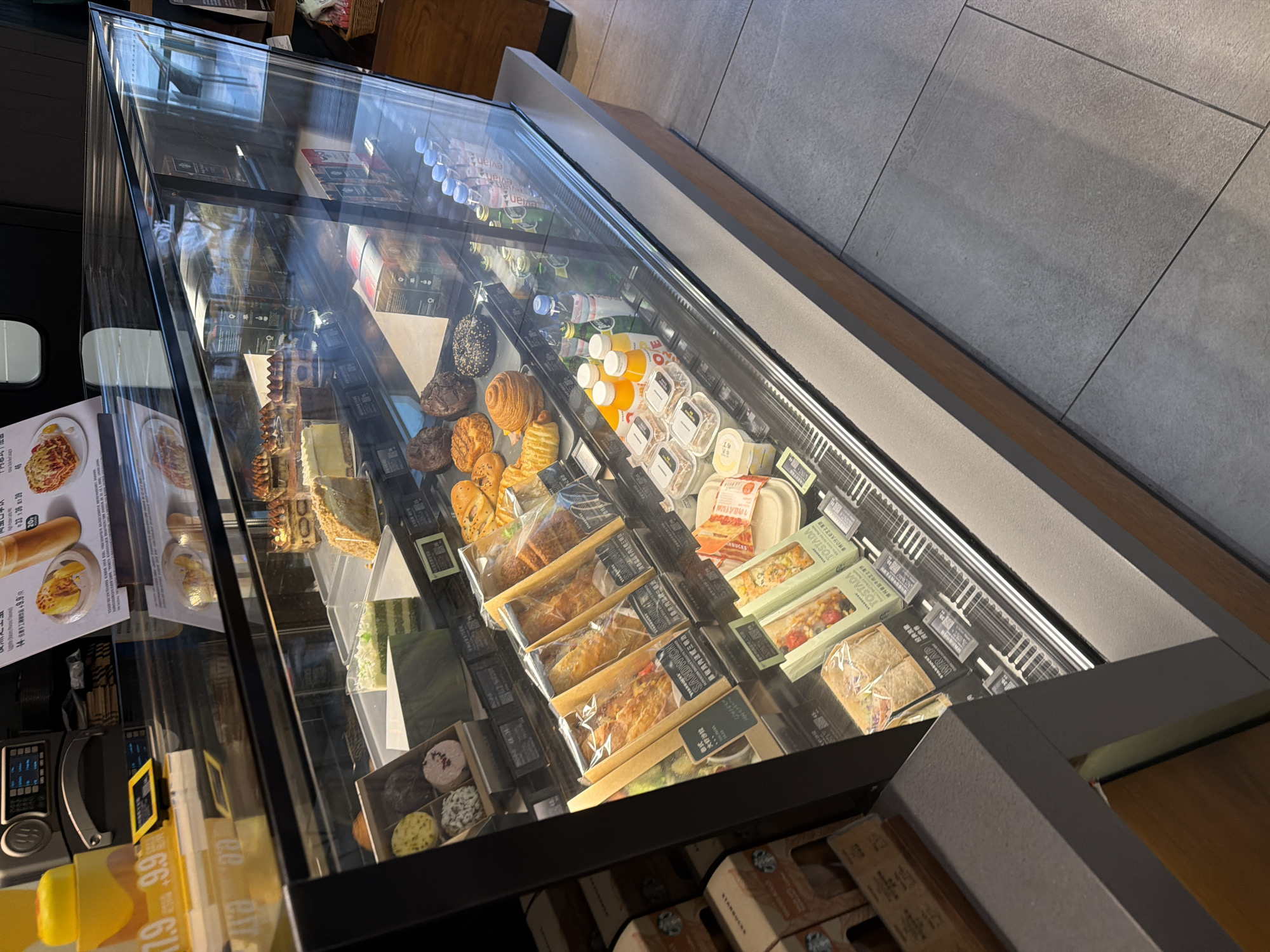 This is a slightly tilted, medium-angle shot of a glass display case filled with various food and beverage items, likely located in a cafe or bakery setting. The case contains a variety of pastries, sandwiches, salads, and drinks, neatly arranged on shelves. The camera angle reveals some of the items from the front and top. Signs and labels are visible, some with Chinese text. The floor is made of large grey tiles, and wooden elements are visible along the edges of the display case. The location may be within the city of Yuhuatai District, China, if that information is needed to provide context.