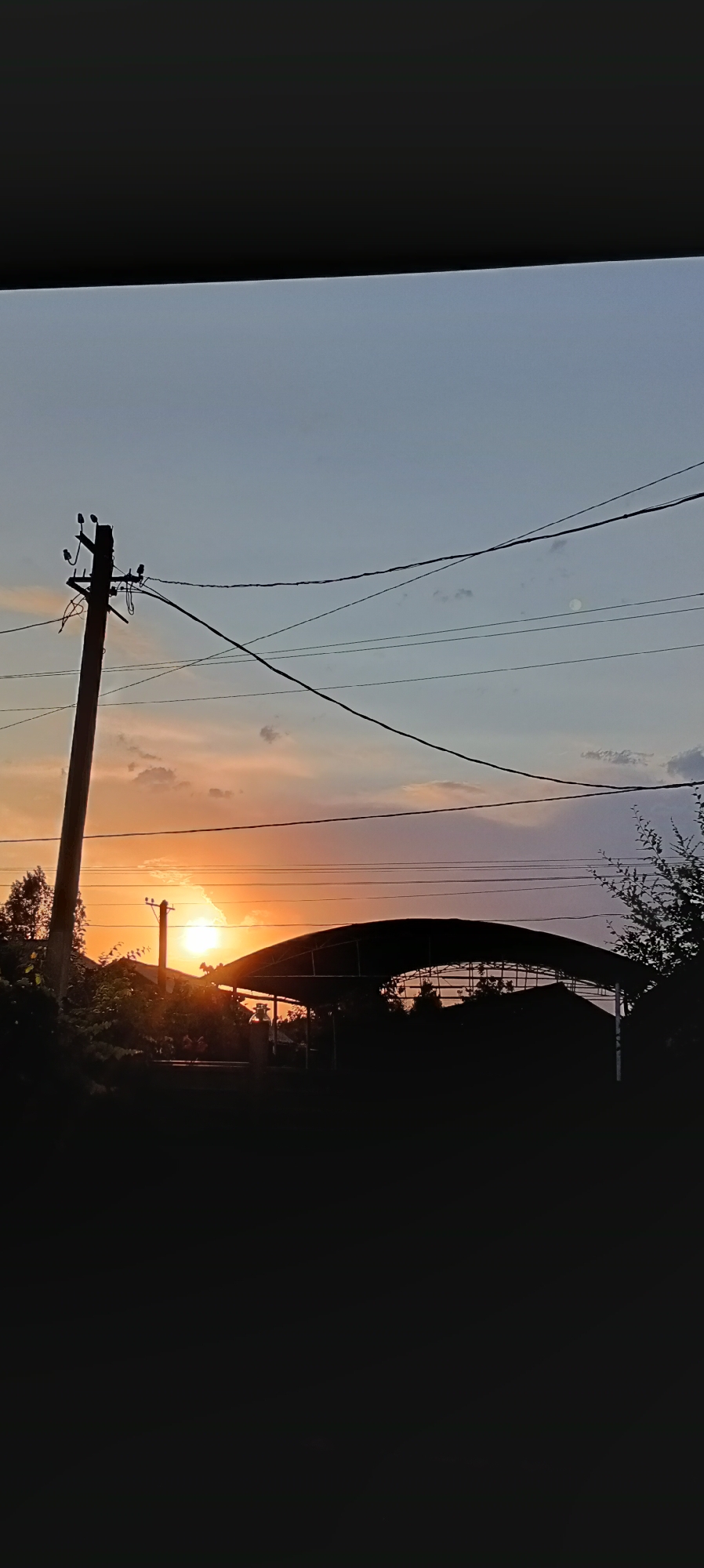 A silhouette of a utility pole with wires against a dusky sky is visible on the left side of the image. In the background, a curved structure, possibly a canopy or roof, is silhouetted against the setting sun, which appears as a bright orange disc low on the horizon. The sky transitions from a warm orange-pink near the sun to a pale blue overhead, with wisps of clouds. Trees and foliage are also visible in silhouette along the lower half of the image. The scene appears to be an outdoor setting, possibly in a rural or semi-urban area.