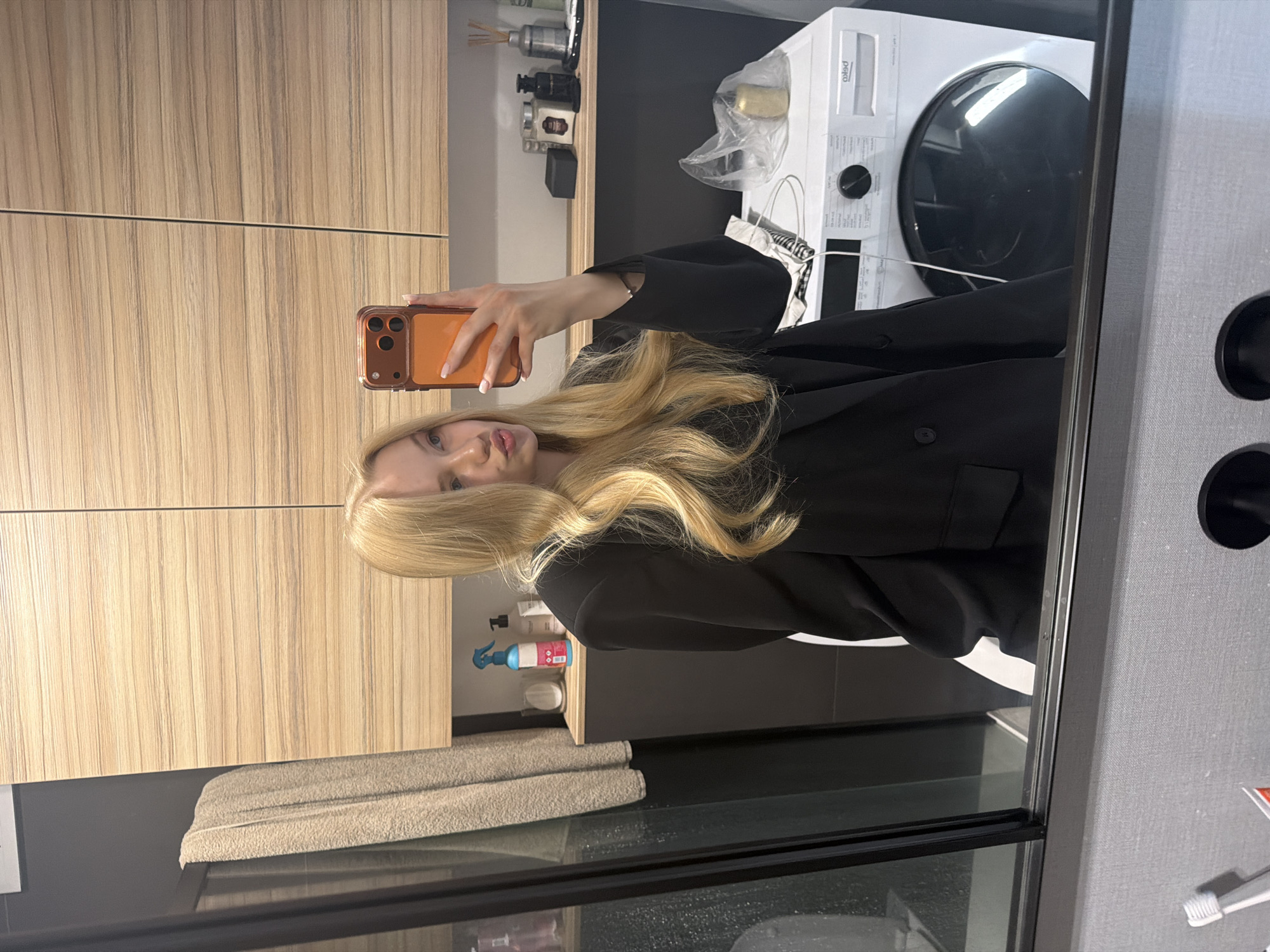 A young woman with long, wavy blonde hair and light-colored nails is featured in a mirror selfie, looking towards the camera with a subtle pout. She is wearing a black blazer or jacket. She holds a smartphone with an orange case to take the photo.
The background, visible through the mirror's reflection, depicts a modern interior, likely a bathroom or utility room. To the left, there are light wooden cabinets with a vertical grain pattern. Above the dark countertop to the left, various bottles, including a blue spray bottle with "BI-PHASE" partially visible text, and diffusers are neatly arranged.
In the upper right, a white front-loading appliance, possibly a dryer or washing machine, is visible with "BEKO" or "Pekto" text on its front, along with other unreadable labels. Below this, two neatly folded light-colored towels rest on a dark surface. To the far right, a gray, textured wall is visible, with a toothbrush in red packaging partially seen. The setting appears clean and organized.