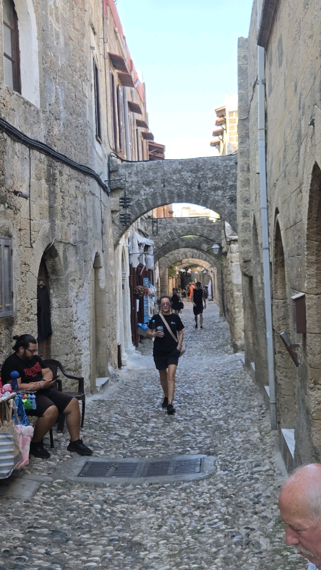 The image depicts a cobblestone street lined with ancient stone buildings, likely within the Municipal Unit of Rhodes, Greece. The architecture suggests a historical setting, with arched passages and rough-hewn stone walls. Sunlight filters through the narrow street, illuminating the scene.
In the foreground, on the left, a man with dark hair and a beard is seated on a dark chair, looking down at his phone. He is wearing a black t-shirt with white lettering and dark shorts. Next to him, a small market stall displays various colorful items, possibly souvenirs.
Further down the street, a woman is walking towards the camera. She is wearing a black t-shirt, black shorts, sunglasses, and a crossbody bag. She is holding a drink in her right hand. Behind her, other figures can be seen walking in the distance, suggesting a public thoroughfare. The street itself is paved with rounded pebbles. The overall impression is a busy, yet timeless, European street scene.
