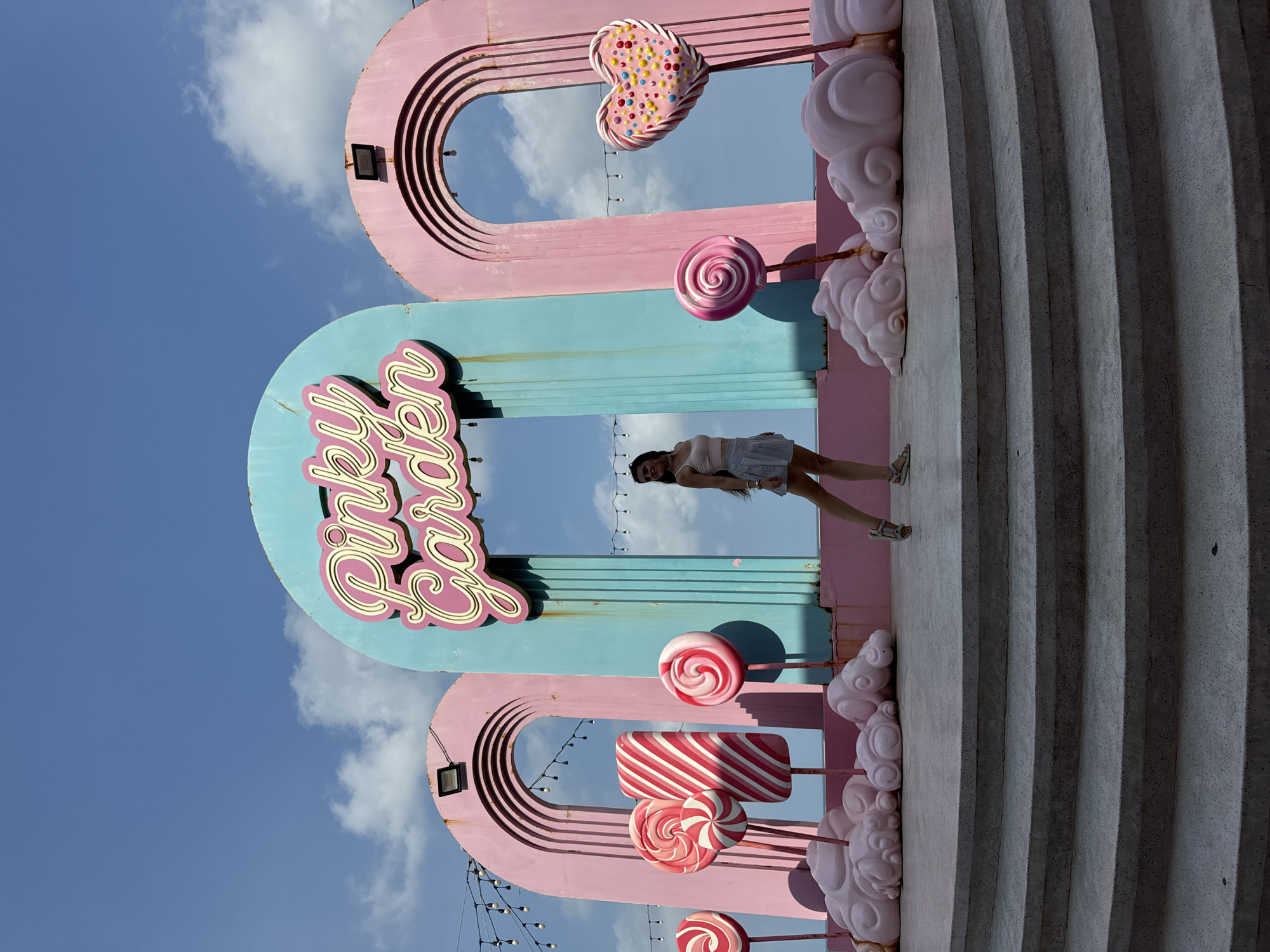 A full-body shot shows a woman posing in front of a whimsical, candy-themed backdrop with the words "Pinky Garden" in large, cursive letters. The backdrop features large, pastel-colored arches and giant lollipop and candy stick props, evoking a playful, sweet atmosphere. The woman is standing on a wide, tiered concrete platform, facing the camera with a slight smile. She is wearing a light pink crop top and a white pleated skirt, with her hair down. The sky above is blue with scattered white clouds, and string lights are visible in the background, suggesting an outdoor setting in the early evening. The scene appears to be located in Tien Thanh Ward, Vietnam, a coastal area known for its picturesque beaches and tourist attractions, which might explain the vibrant, decorative setup.