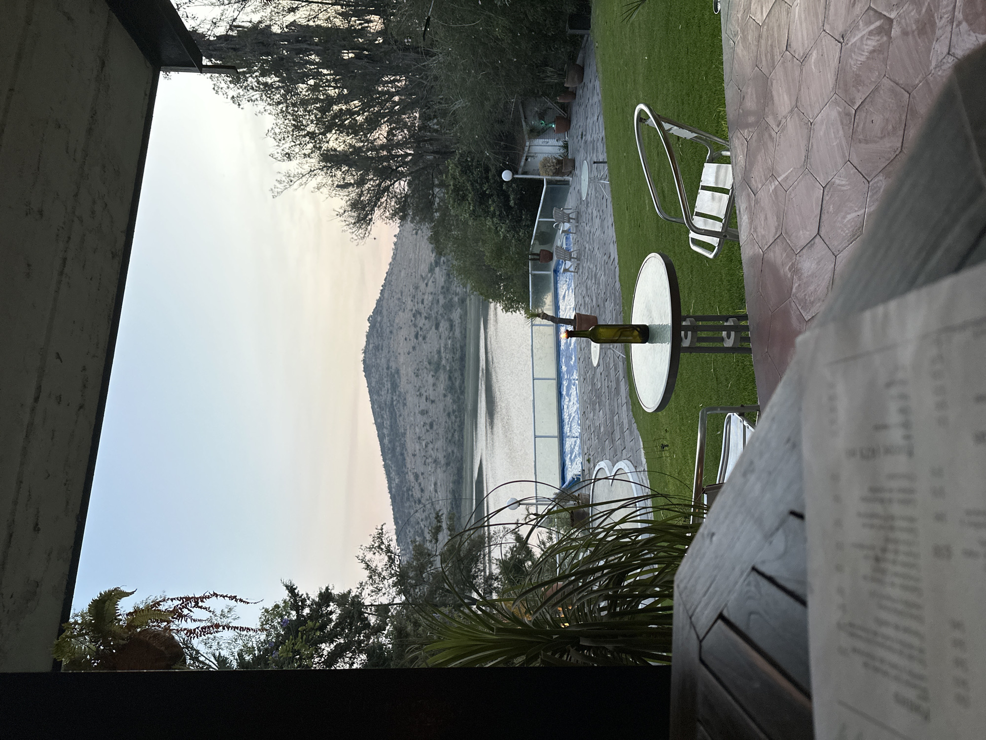 From an elevated position, the image captures a tranquil scene overlooking a lake and a distant mountain. The view is framed by a structure on the left, partially obscuring the sky, and a table with a menu on the right. The foreground features outdoor seating with modern metal chairs around small, round tables. A wine bottle is positioned on one table. Beyond the seating area, a well-maintained green lawn leads to a terrace overlooking the lake. The lake's edge is defined by a white fence and a blue-tiled pool area. The background is dominated by a hill or mountain range under a muted sky. The image gives the impression of an outdoor restaurant or resort in El Oasis Valsequillo, Mexico.