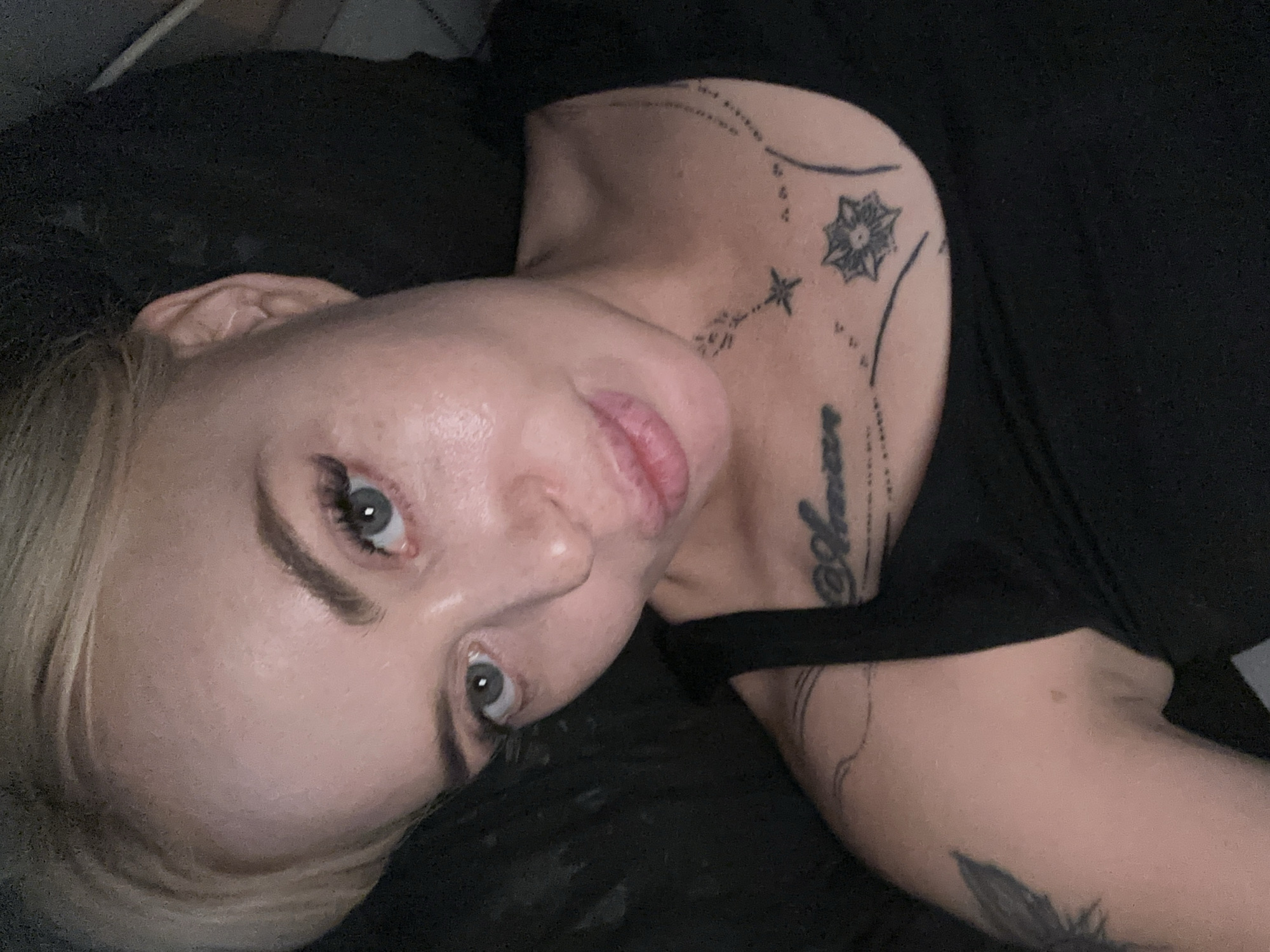 This media file shows a close-up, overhead shot of a person with blonde hair lying down. The person is wearing a black t-shirt, and part of their chest and shoulder are visible. They have several tattoos on their neck and arm, including what appears to be a star or flower design and some script. The lighting suggests it may be indoors, possibly at night or in a dimly lit environment, as there are no strong shadows or signs of natural light. The person's eyes are open and looking towards the camera, and their lips are slightly parted. The overall mood is intimate and personal, with the subject's gaze being direct and engaging. There is no discernible text in the image.