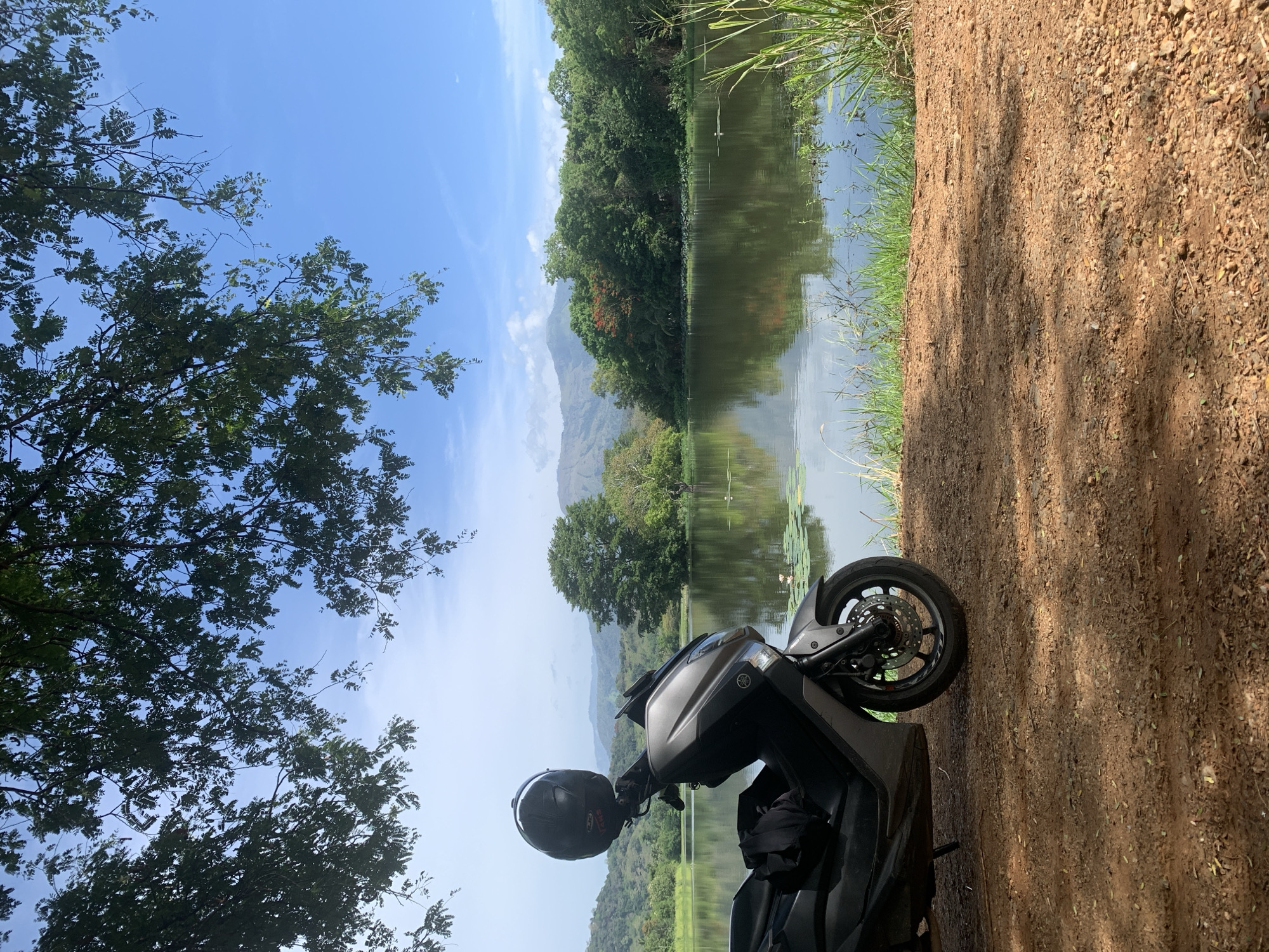A dark grey scooter is parked on a dirt path next to a calm lake. A black helmet rests on the handlebars of the scooter, and a dark fabric item is draped over the seat. The lake is surrounded by lush green trees and rolling hills in the distance. The sky is a clear blue with a few scattered clouds. The overall scene suggests a peaceful, natural setting, likely in a rural area. The location is the city of Buduruwagala, Sri Lanka.