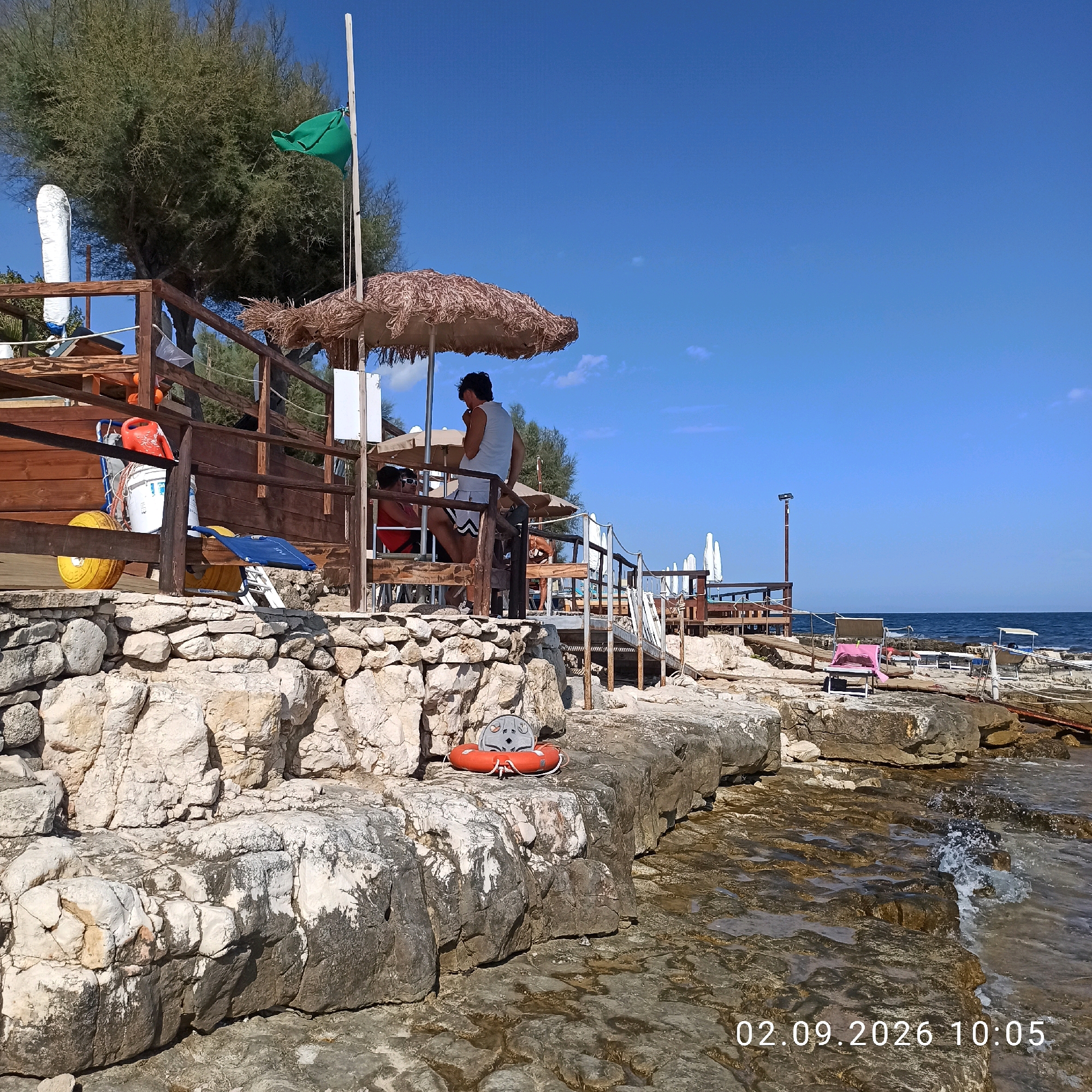 A sunny day is depicted at a rocky coastal area in Molfetta, Italy, near a lifeguard station. The foreground features large, weathered rocks with the sea lapping at them. A red life preserver with a grey seat is placed on the rocks. Behind the rocks, a wooden structure, likely a lifeguard station or beach bar, is visible. It has a thatched umbrella and a wooden railing. Several people are present at the structure. One person, wearing a white tank top and shorts, stands looking down. Another person in a red shirt and sunglasses is seated. Further back, a person in a swimsuit is bending over. Various objects are stored around the structure, including orange watering cans, a white bucket, a yellow wheel, and beach chairs. In the background, the blue sea stretches to the horizon under a clear blue sky. A green flag flies from a pole near a tree. Several white beach umbrellas are set up in the distance. The date stamp "02.09.2026 10:05" is visible in the lower right corner.