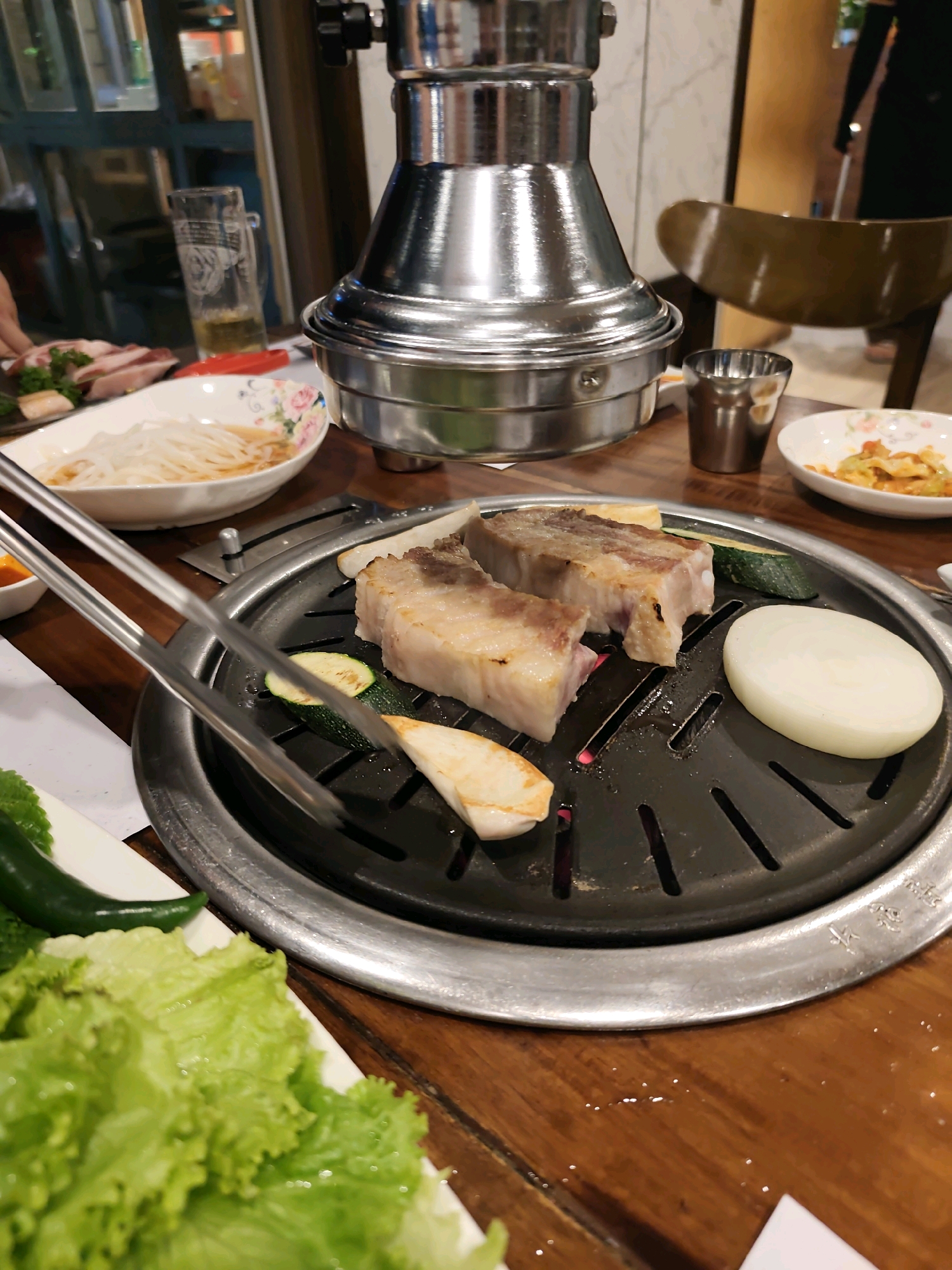 This is a close-up, medium-shot, eye-level photograph of a Korean barbecue grill set up on a wooden table. In the foreground, a circular grill with a black, perforated surface is centrally located. Two thick, rectangular pieces of raw meat, likely pork belly, are being cooked on the grill, with visible char marks and some fat rendering. Sliced zucchini, a piece of daikon radish, and a thick slice of white onion are also present on the grill. To the left of the grill, a pair of silver metal tongs is positioned, with its tips gripping a piece of meat. In the lower left of the frame, a large cluster of bright green lettuce leaves is visible, along with a long, dark green jalapeño pepper. In the background, a tall, silver metal exhaust hood is suspended over the grill. To the left of the hood, a clear glass mug filled with a yellowish liquid, possibly beer, sits on the table. Further left, a white ceramic bowl with a floral pattern contains thinly sliced radishes in a light broth. To the right of the grill, a small, metallic cup and a white ceramic dish with a small portion of colorful food, likely a side dish or banchan, are visible. The table surface is a polished dark wood, showing some light reflections. The overall environment appears to be an indoor restaurant setting.