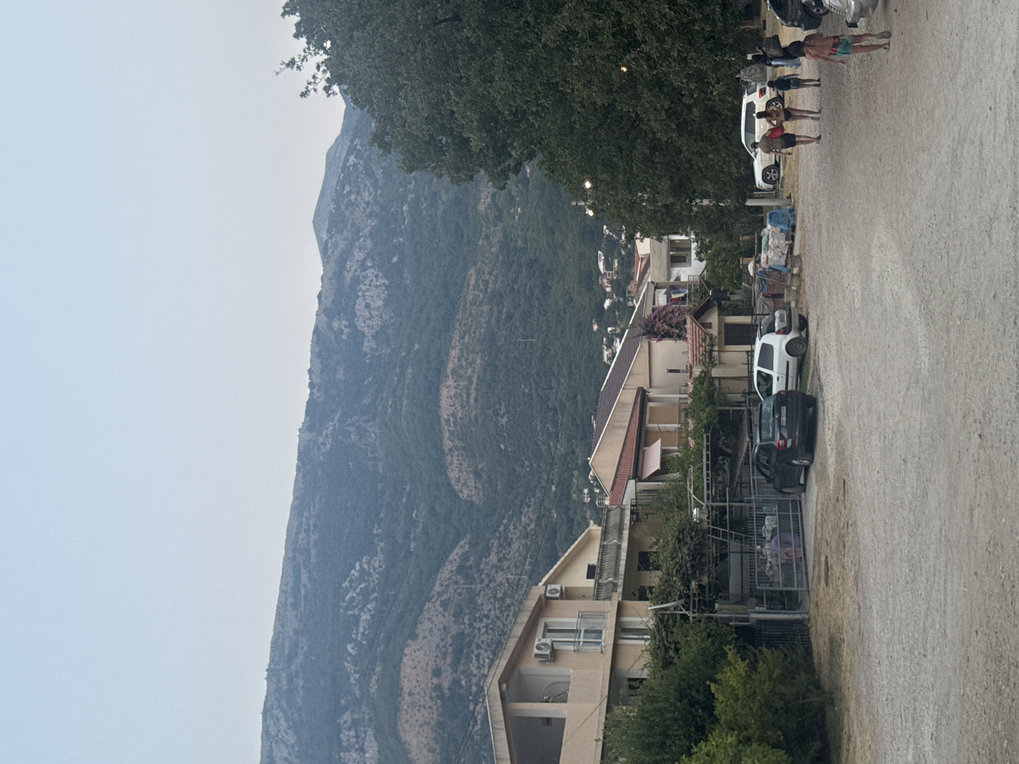 The image depicts a street scene in Sutomore, Montenegro, characterized by a dirt road in the foreground and buildings lining the street. In the background, a large, forested mountain range dominates the skyline. Several vehicles, including cars and a white SUV, are parked along the road. A group of people, including adults and children, are walking on the right side of the road. The architecture of the buildings appears to be residential, with balconies and windows visible. Lush green trees and vegetation surround the buildings and add to the natural beauty of the scene. The sky is a pale blue, indicating it is daytime.