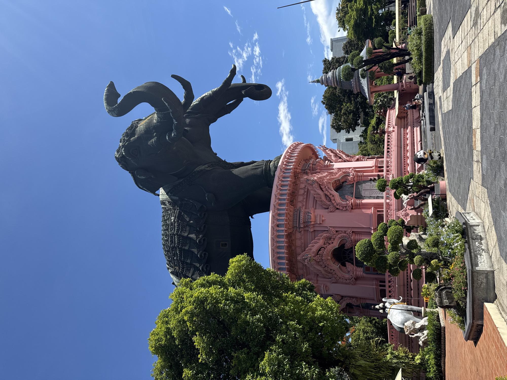 The image showcases a monumental, multi-headed elephant statue, deeply shadowed, perched atop a pink, ornate, multi-tiered structure. This structure features intricate architectural details. The scene is set against a bright blue sky with wisps of clouds. The statue is surrounded by trees. To the right, the edge of a pavement or walkway is visible, along with other architectural elements. The image is taken at the Erawan Museum in Samrong Nuea Subdistrict Municipality, Thailand. Some people appear to be present but are small and indistinct.