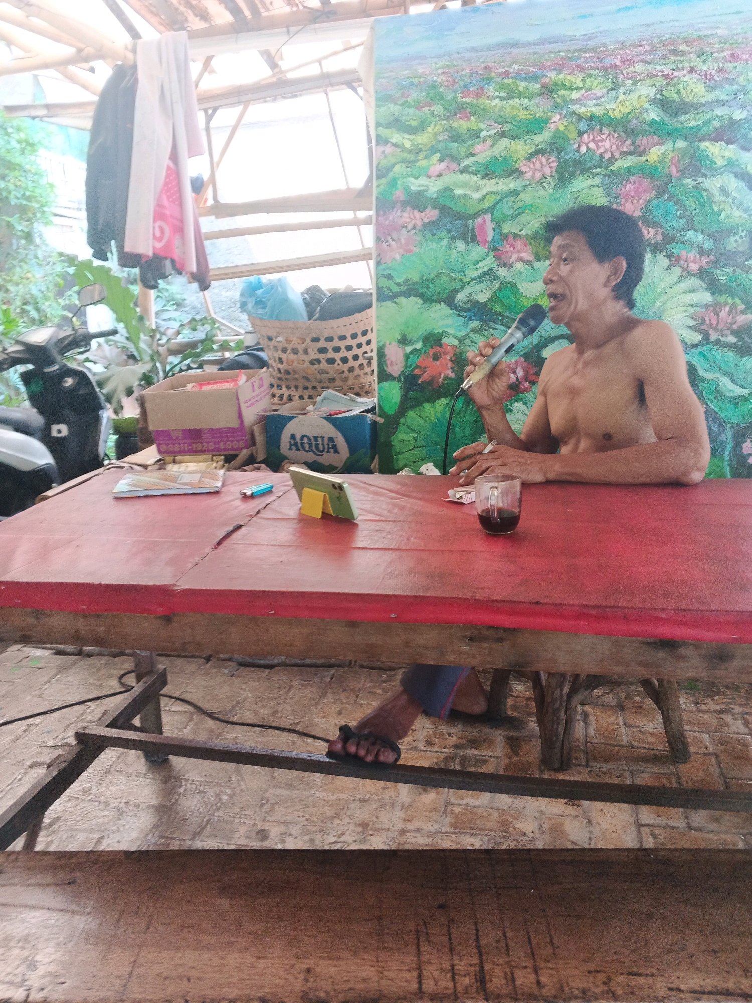 In Ungaran, Indonesia, an elderly East Asian man with short dark hair and a bare chest sits at a red-topped table. He holds a microphone in his right hand and appears to be speaking or singing. To his left, a small glass of dark liquid sits on the table. Behind the man, a large, colorful painting depicts a landscape filled with pink lotus flowers and green lily pads. The table is covered with various items, including a cigarette pack, a lighter, a mobile phone resting on a yellow stand, and a small notebook. In the background, a motorcycle is parked to the left, and laundry hangs from a wooden structure overhead. A woven basket and cardboard boxes are also visible in the background, suggesting a somewhat cluttered outdoor or semi-outdoor environment. The man's feet, clad in dark flip-flops, are visible resting on a metal bar beneath the table.