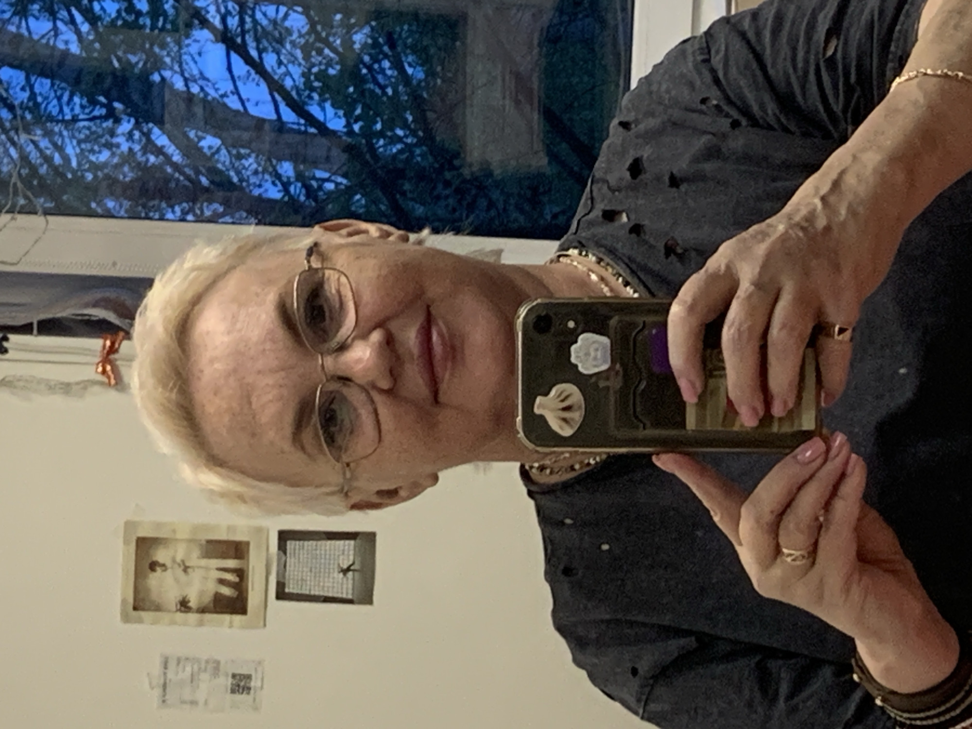 A woman with short blonde hair and glasses is taking a selfie with her phone. She is wearing a dark gray shirt with holes and has a gold chain around her neck. She also has multiple bracelets on her wrist and rings on her fingers. The background shows a window with tree branches visible outside. The interior wall features two framed pictures and some smaller paper items. The location cannot be determined to be Chaikovsky, Russia based on the visual data provided.