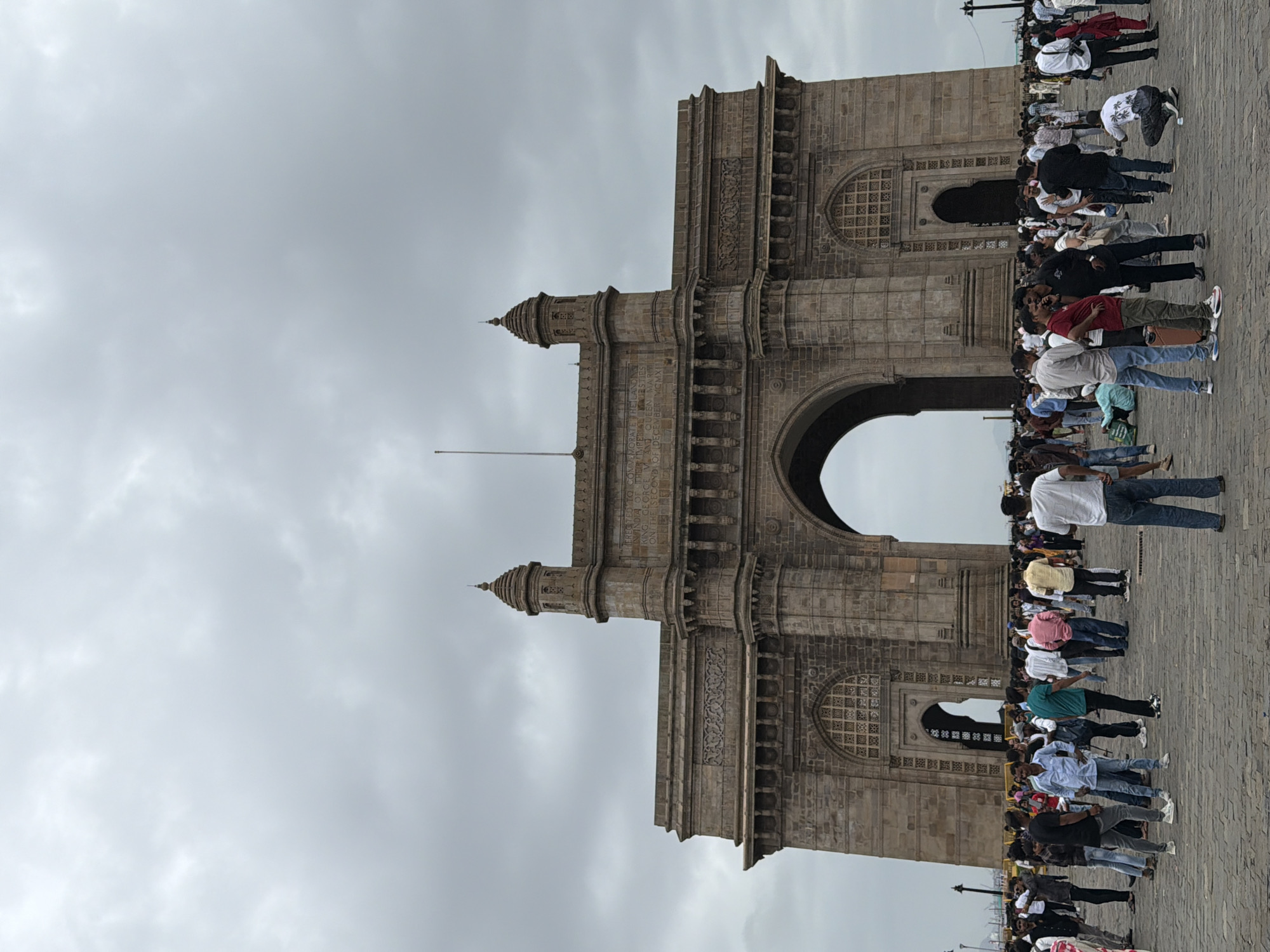 The image shows a wide shot of the Gateway of India, a monument in Mumbai, India, under a cloudy sky. A large crowd of people are gathered in front of the monument, many of them appearing to be tourists. The monument is made of stone and features a large archway. The ground in front of the monument is paved with stone. The overall scene is one of a busy public space with a prominent historical landmark.
