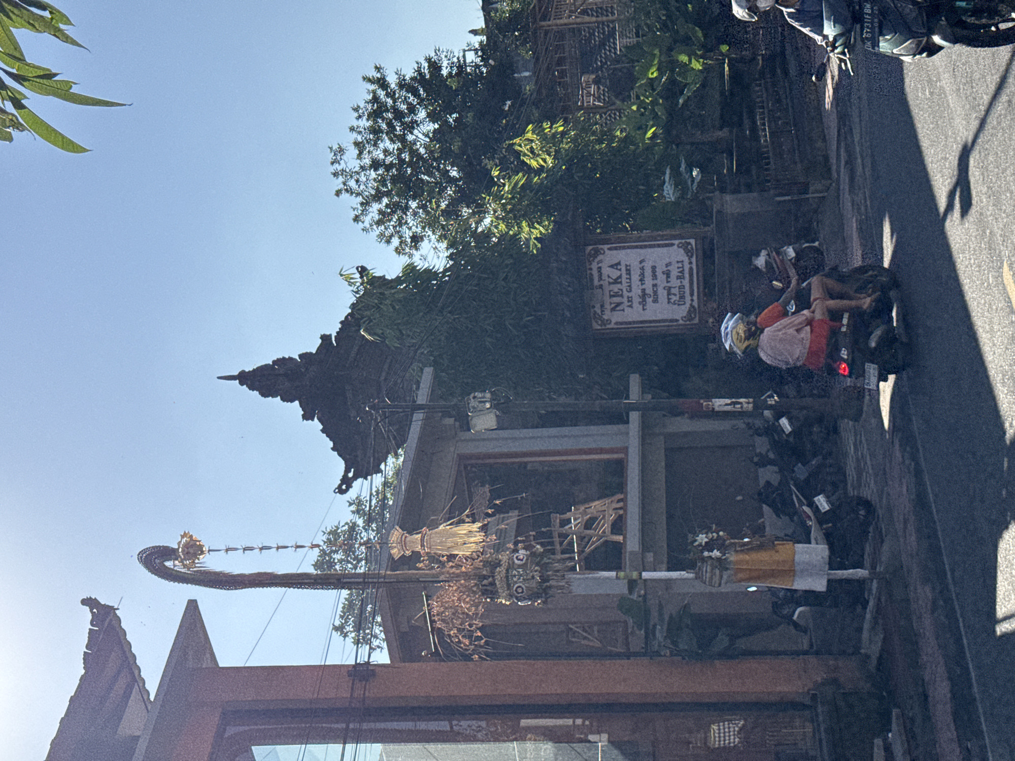 In a street scene in Ubud District, Indonesia, under a clear blue sky, the sun casts strong shadows. To the left, a modern building with a red facade and large windows stands. A traditional Balinese decorative item, likely a PENJOR, hangs from a pole in front of the building. Further back, a tiered Balinese temple structure is visible amongst lush green trees. A sign for "NEKA ART GALLERY SINCE 1909 UBUD-BALI" is mounted on a wall. Several motorbikes are parked along the street, and two people are riding a motorbike in the foreground, with another person on a motorbike to the right. The street is paved and appears to be a busy thoroughfare.