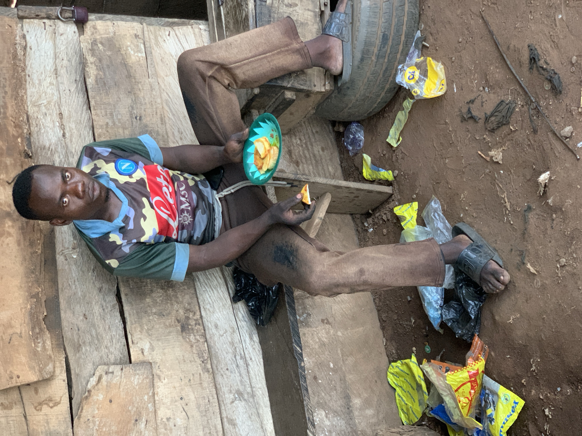 In Gbeleko, Nigeria, a dark-skinned man is sitting outdoors, possibly on or near a wooden platform, appearing to eat. He's wearing a multicolored camouflage t-shirt with "Napoli" written on it, brown pants, and black sandals. He is holding a green plastic plate with food and a piece of orange fruit in his hand. His right foot rests on a tire. Litter, including yellow plastic bags, is scattered on the ground. The ground appears to be dirt and mud.
