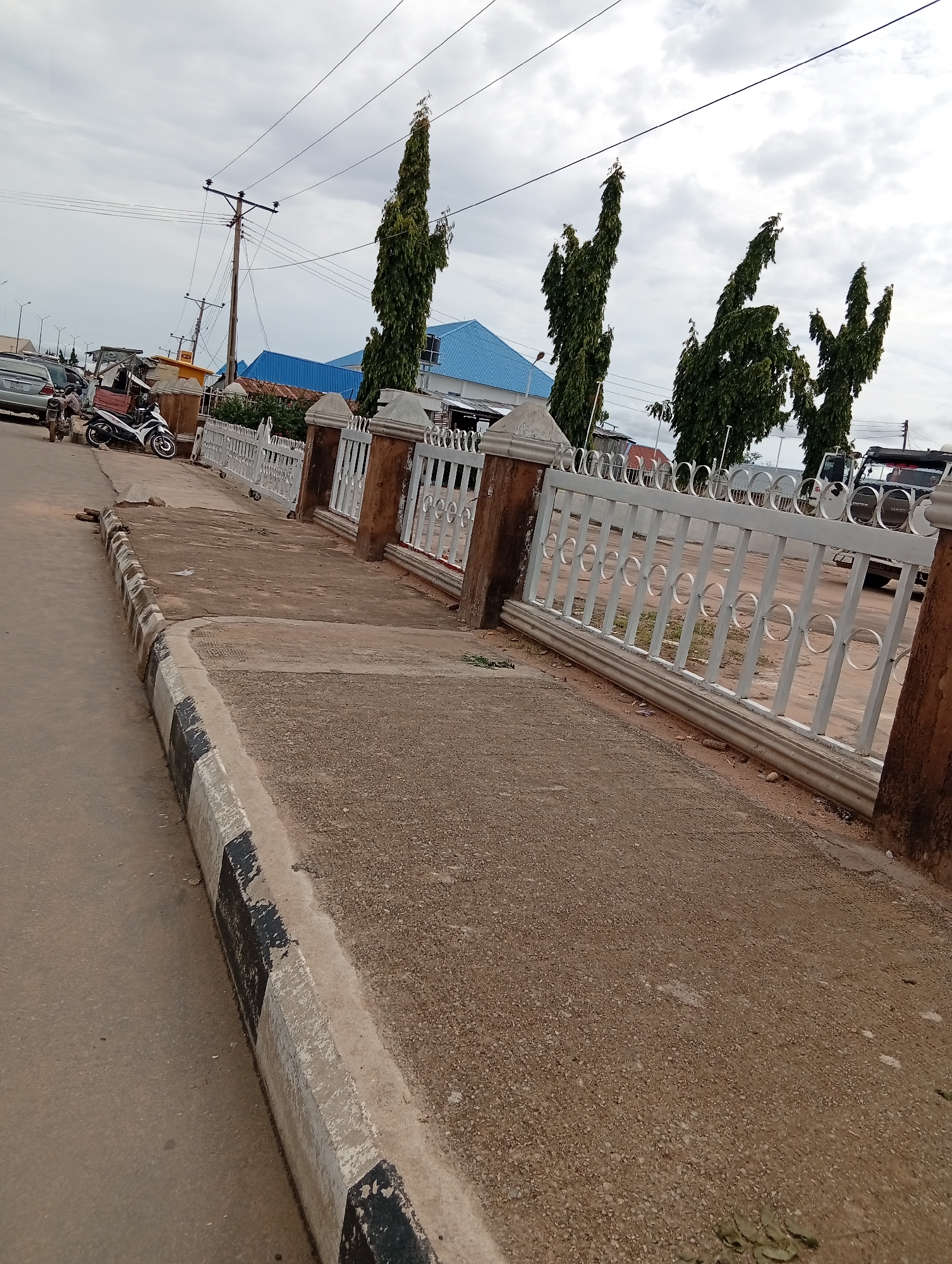 The image depicts an outdoor scene in Jalingo, Nigeria, on a cloudy day. A paved walkway runs alongside a road, bordered by a decorative white fence with circular accents. Beyond the fence, several tall, slender trees with dense foliage stand against the overcast sky. A building with a distinctive blue roof is visible in the background, along with utility poles and power lines.
On the road to the left, a motorcycle is parked, and a person appears to be seated on it. Several other vehicles are also present, including what looks like a silver car and a darker SUV or truck. The road itself is made of asphalt, and the walkway is composed of concrete with a gravelly surface. The curb separating the road from the walkway is painted with alternating black and white stripes, showing signs of wear.
The overall impression is of a street scene in a town or city, likely during daytime given the ambient light, though the heavy cloud cover suggests it might be overcast. There is no discernible text visible in the image. The scene appears quiet, with no overt activity or people prominently featured beyond those near the parked motorcycle.