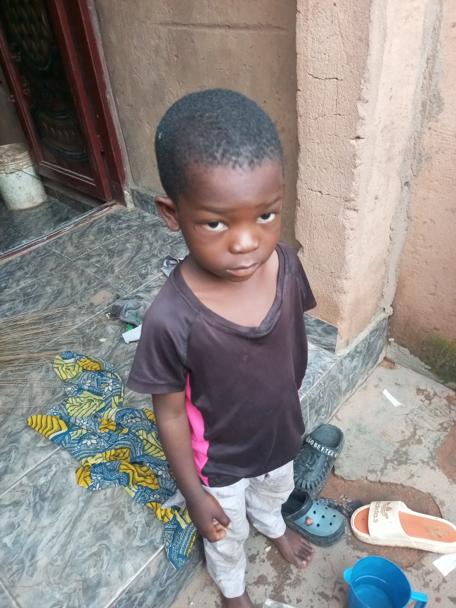 An outdoor, ground-level, medium-shot photograph captures a young Black child standing on a tiled surface in front of a textured, earth-toned wall. The child, appearing to be around four or five years old, faces the camera with a neutral expression. They are wearing a dark t-shirt with pink accents on the sleeves and light-colored shorts.
Scattered on the ground around the child are various items. To the child's left lies a folded piece of fabric with a vibrant, patterned design featuring yellow, blue, and white motifs. Next to this fabric is a broom. To the child's right, several pairs of footwear are visible: a pair of black clogs with white lettering that reads "GO BETTER," a light blue clog, and a white slide sandal with an orange sole and an Adidas logo. A blue plastic pitcher, partially filled with water, sits on the ground in the foreground.
The environment appears to be a residential area. To the far left of the frame, a portion of a dark red door and a white bucket are visible. The wall behind the child exhibits a rough, stucco-like texture with visible cracks and some green moss or algae growth at the lower right edge. The tiled ground is grey with a marbled pattern. The overall scene suggests a simple, outdoor living space. The location is identified as Tempure, Nigeria.