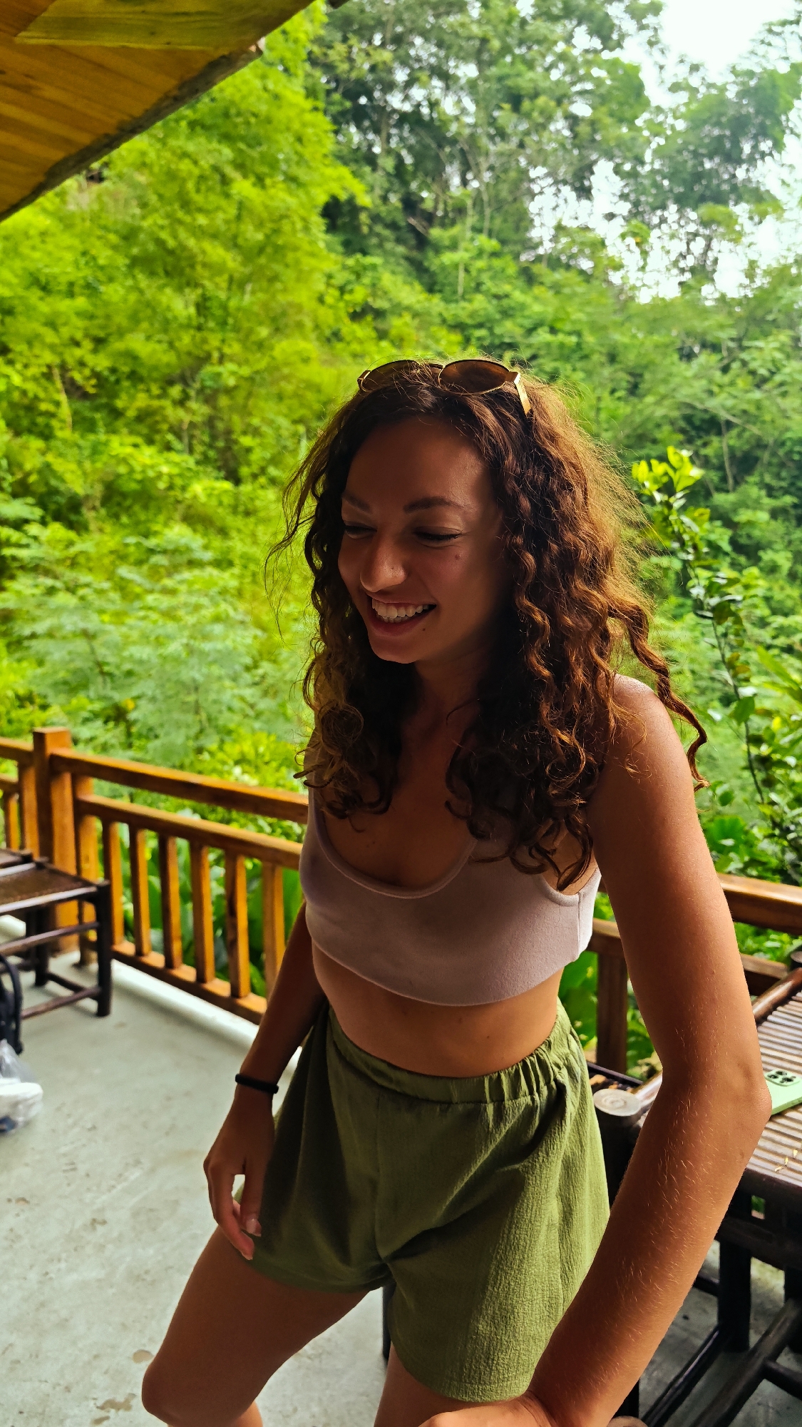 A young woman with curly brown hair and sunglasses pushed onto her head is smiling and looking down. She is wearing a light pink crop top and olive green shorts. She stands on a wooden porch or balcony with a railing. In the background, lush green trees and foliage are visible, suggesting a tropical or jungle setting. The floor of the porch appears to be concrete. A wooden table and chairs are partially visible to the left of the woman. To the right, another wooden table is present. The overall impression is a relaxed, outdoor scene, likely in a natural environment. The location is described as Pù Luông Commune, Vietnam.