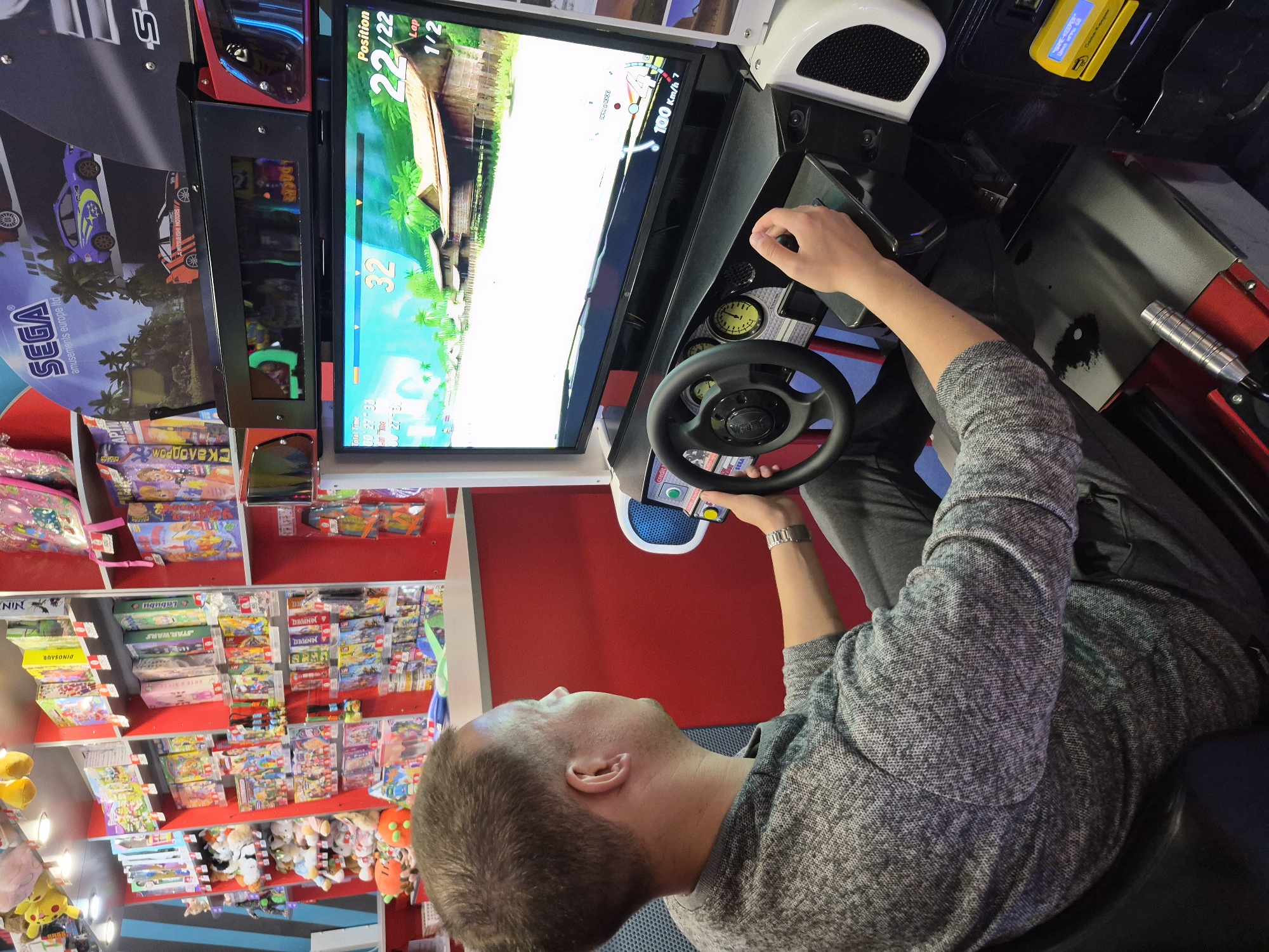 The image shows a man playing a racing arcade game. He's gripping the steering wheel and looking intently at the screen, which displays the game's interface showing his position, lap number, and speed. The arcade machine has a gear shift and other controls visible. Behind the man, shelves are filled with various toys and board games. The game advertises the name "SEGA". The OCR detected the word "Скалодром" which means "climbing wall" in Russian. The full text in the first column of the shelf reads "Labubu, STARWARS, DINOSAUR, MINIVELL". The text on the screen reads "Position 22/22, Lap 1/2". The location in the city of Tobolsk, Russia is not known.