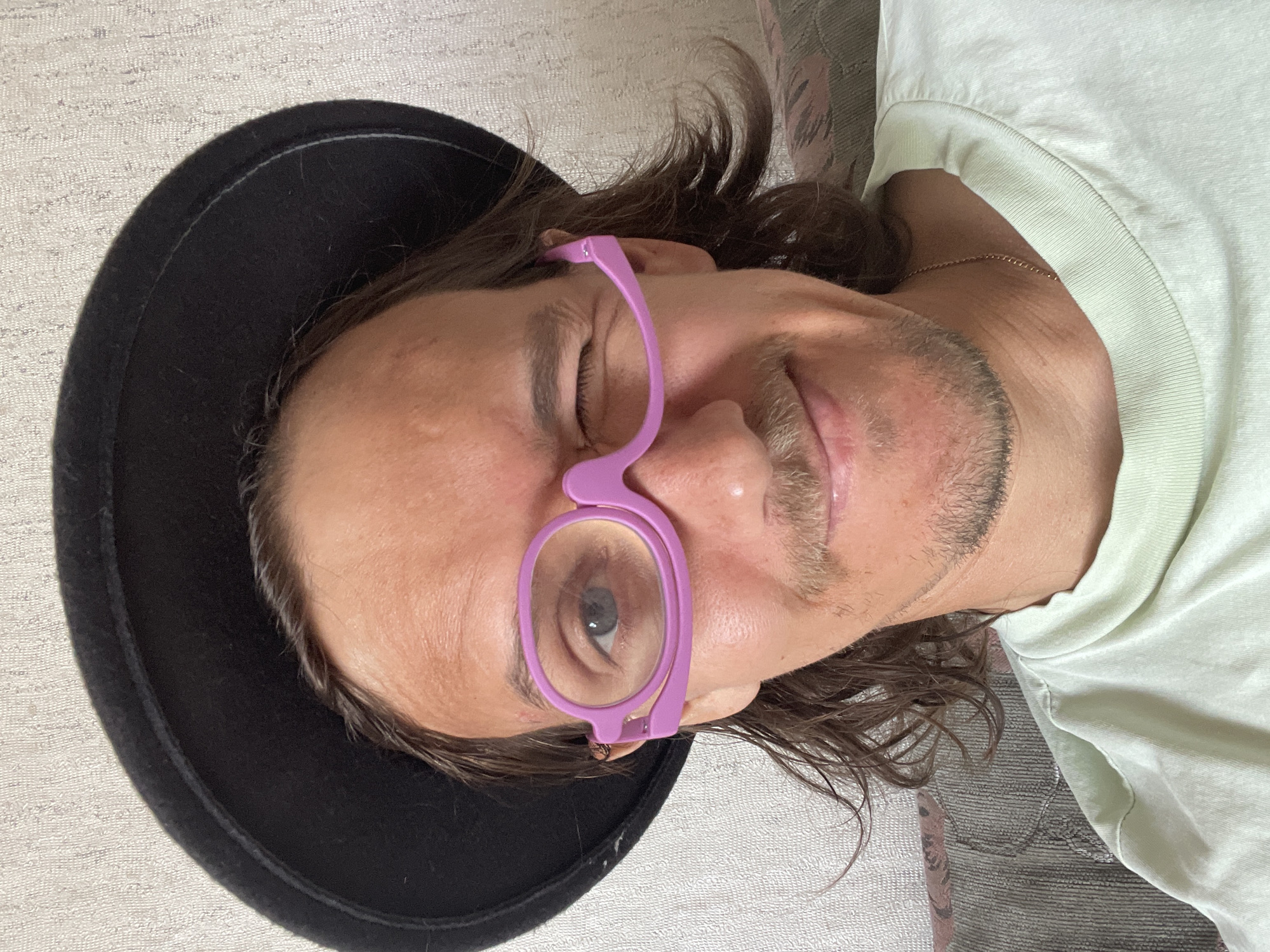 A person wearing a black hat and lavender glasses is lying down. The person is wearing a light green t-shirt. The location is Перевозное, Russia.