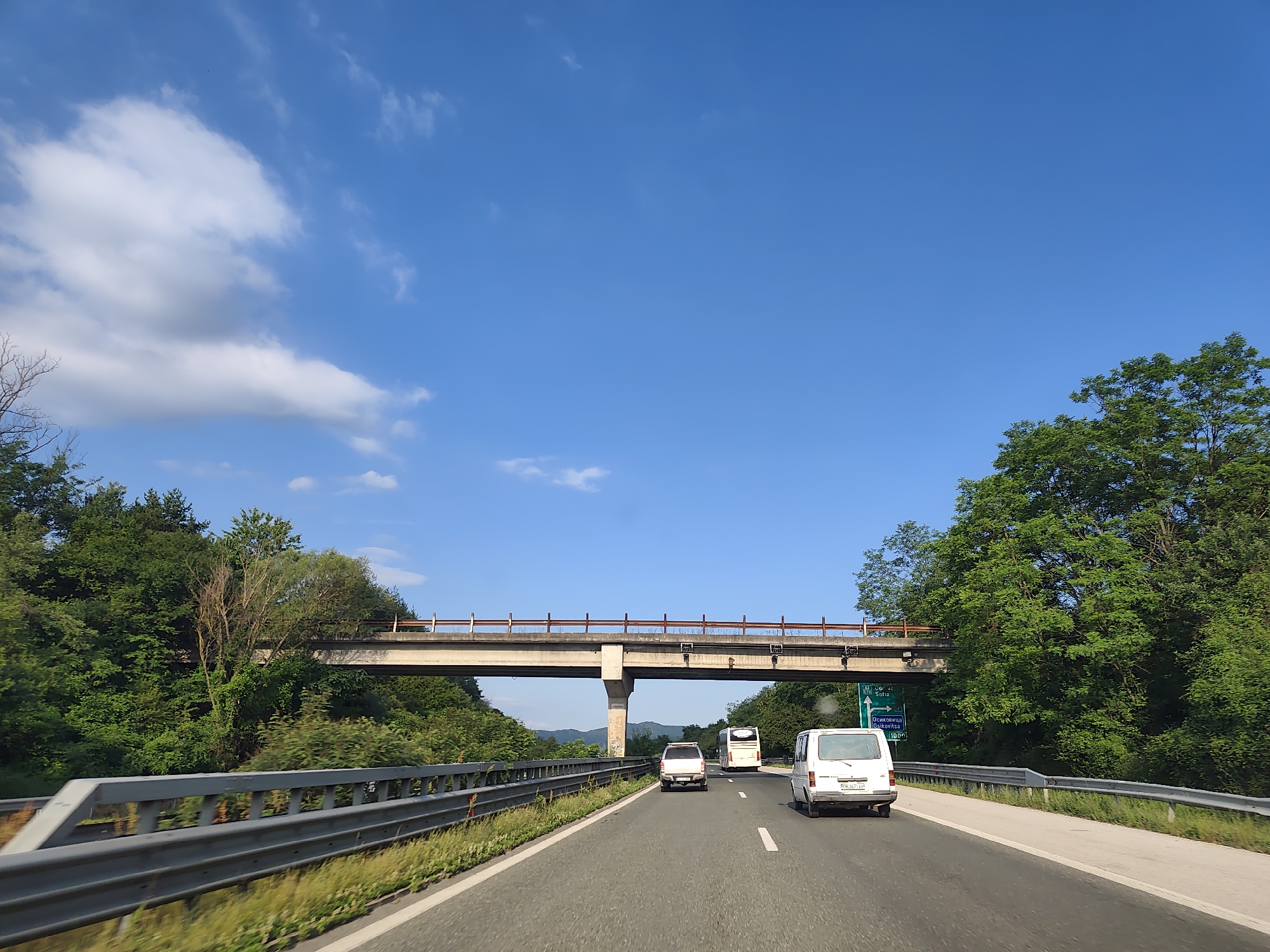 On a bright, sunny day with a clear blue sky and scattered white clouds, a multi-lane highway stretches forward. The road is lined with metal guardrails on both sides, and lush green trees and foliage flank the highway. An elevated concrete overpass, supported by a single large pillar, crosses the highway. On the highway, several vehicles are visible: a white van in the foreground, followed by a white SUV, and a white bus further ahead. A green road sign with white text is positioned on the right side of the highway, indicating directions to Osikovitsa and Sofia, with a distance of 100 km. The scene suggests a typical day of travel on a highway outside of a city.