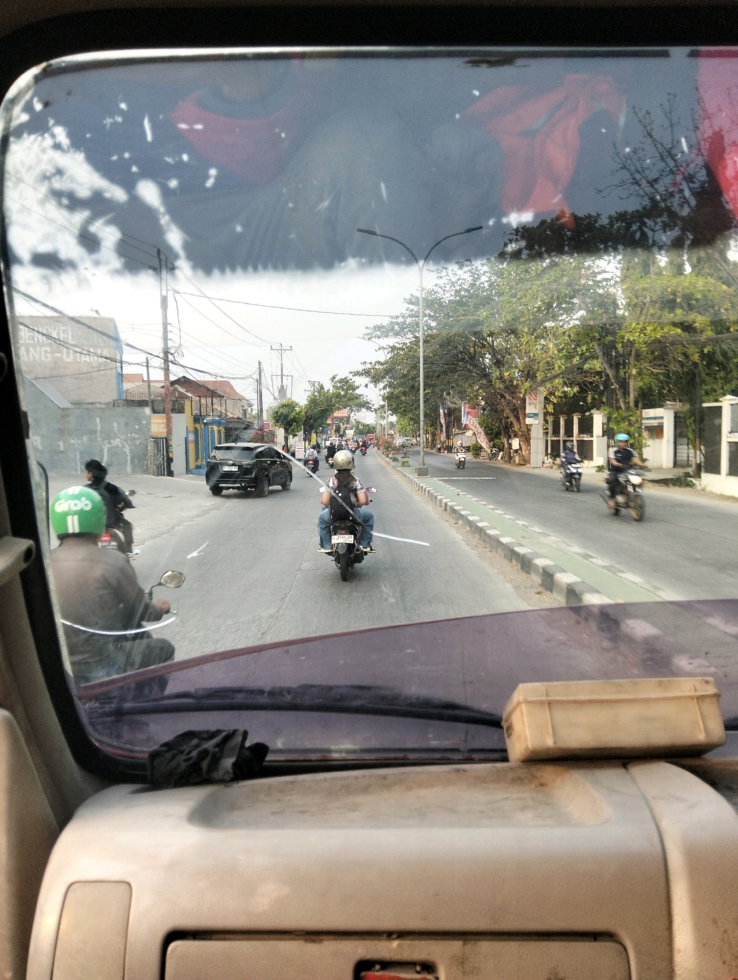 The image, taken from inside a vehicle, shows a view through the windshield of a street scene in Semarang, Indonesia. Several people are riding motorcycles, some in the foreground and others further down the road. In the foreground, a person wearing a green helmet with the "Grob" logo is riding a motorcycle, their back to the camera. To the right, another person on a motorcycle is also visible from behind, wearing a light-colored helmet and jeans. A black SUV is seen driving in the same direction on the road. The street is lined with trees and buildings, including what appears to be a shop with signage. Overhead power lines are visible, common in Indonesian cityscapes. Reflections on the windshield show a blurry red and dark object, possibly clothing or fabric, and the sky. The dashboard of the vehicle is visible in the lower part of the image, with a small beige box resting on it. The overall lighting suggests it is daytime.