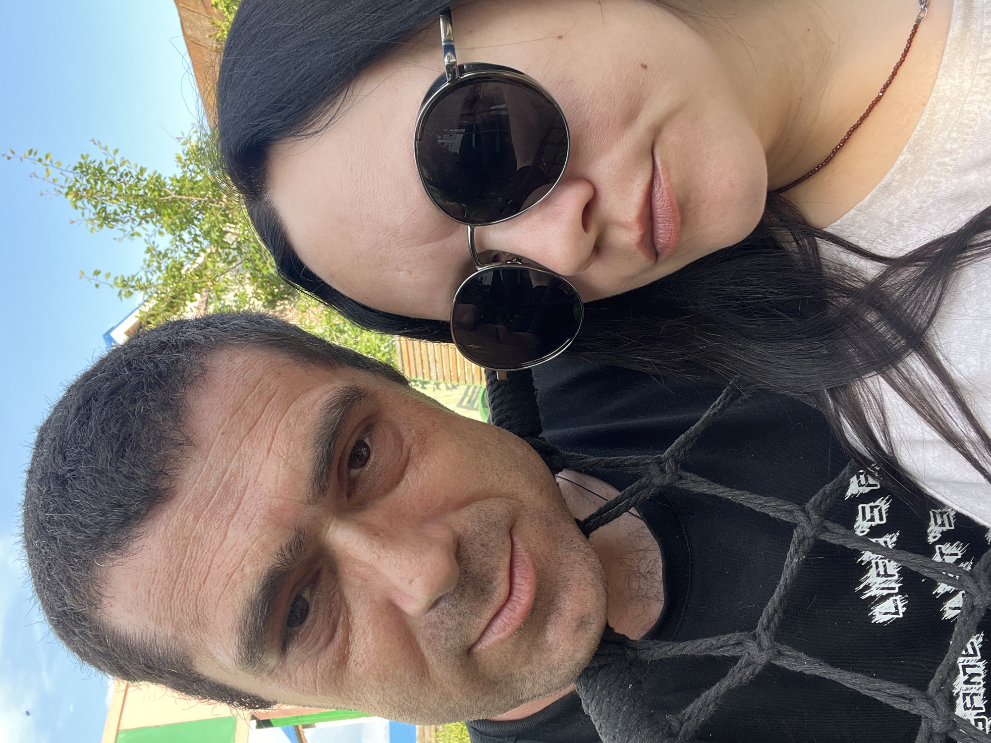 Two people, a man and a woman, are in the foreground of the image, taking a selfie outdoors. The man, on the left, has dark hair and is looking at the camera with a slight smile. He is wearing a black t-shirt with white graphics, partially obscured by a black net. The woman, on the right, has long dark hair and is wearing large round sunglasses and a light-colored t-shirt. She is also looking at the camera. In the background, there is greenery, a blue sky, and parts of buildings. The scene appears to be in a residential or recreational outdoor area, possibly a backyard, given the presence of a tree and structures.