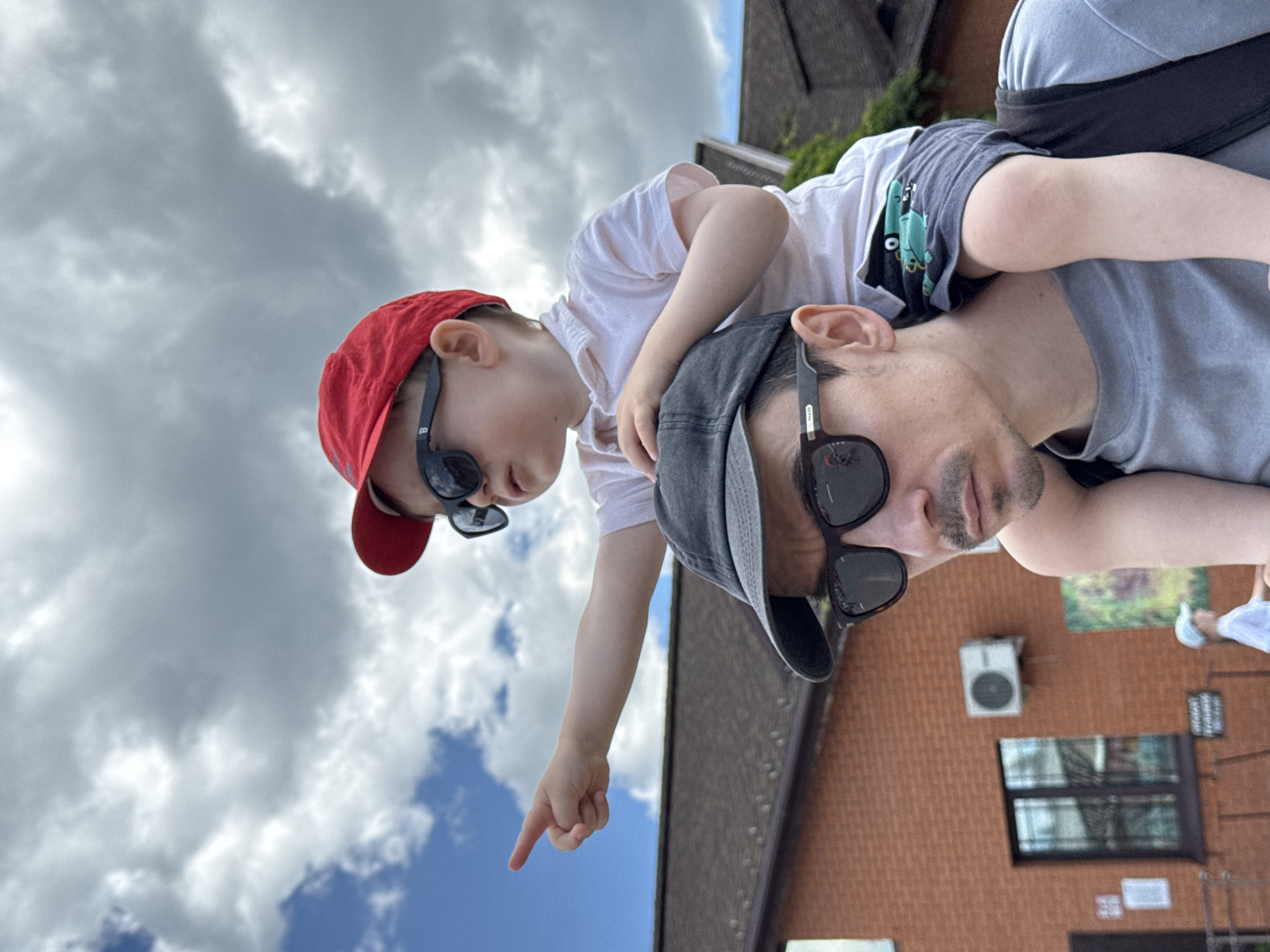 An adult male is carrying a young child on his shoulders in an outdoor daytime setting in Izhevsk, Russia. The adult, appearing to be in his late 20s to 40s, is positioned in the lower right and center of the frame, viewed from the chest up. He wears a grey t-shirt, a dark baseball cap, and dark-rimmed sunglasses, and has a mustache and a short beard. The child, a young boy estimated to be 2-4 years old, sits on the adult's shoulders, wearing a white short-sleeved polo shirt, dark shorts with a green cartoon frog design, a red baseball cap, and dark sunglasses. The child points his right index finger directly to the left side of the frame. The adult's gaze is directed forward and slightly downward.
The background features a red brick building with a dark brown pitched roof. A dark-framed window with security bars and an external air conditioning unit are visible on the brick wall to the left of the subjects. Several white notices or papers are affixed to the wall near the window, and a colorful poster or mural is visible to the right of the AC unit. Green foliage is present on the far right background, partially obscured by the subjects. A blurred partial figure of another individual in a light blue cap and white shirt is seen in the far left background, behind the brick building. The sky is partly cloudy, displaying both blue patches and large, textured grey and white clouds.