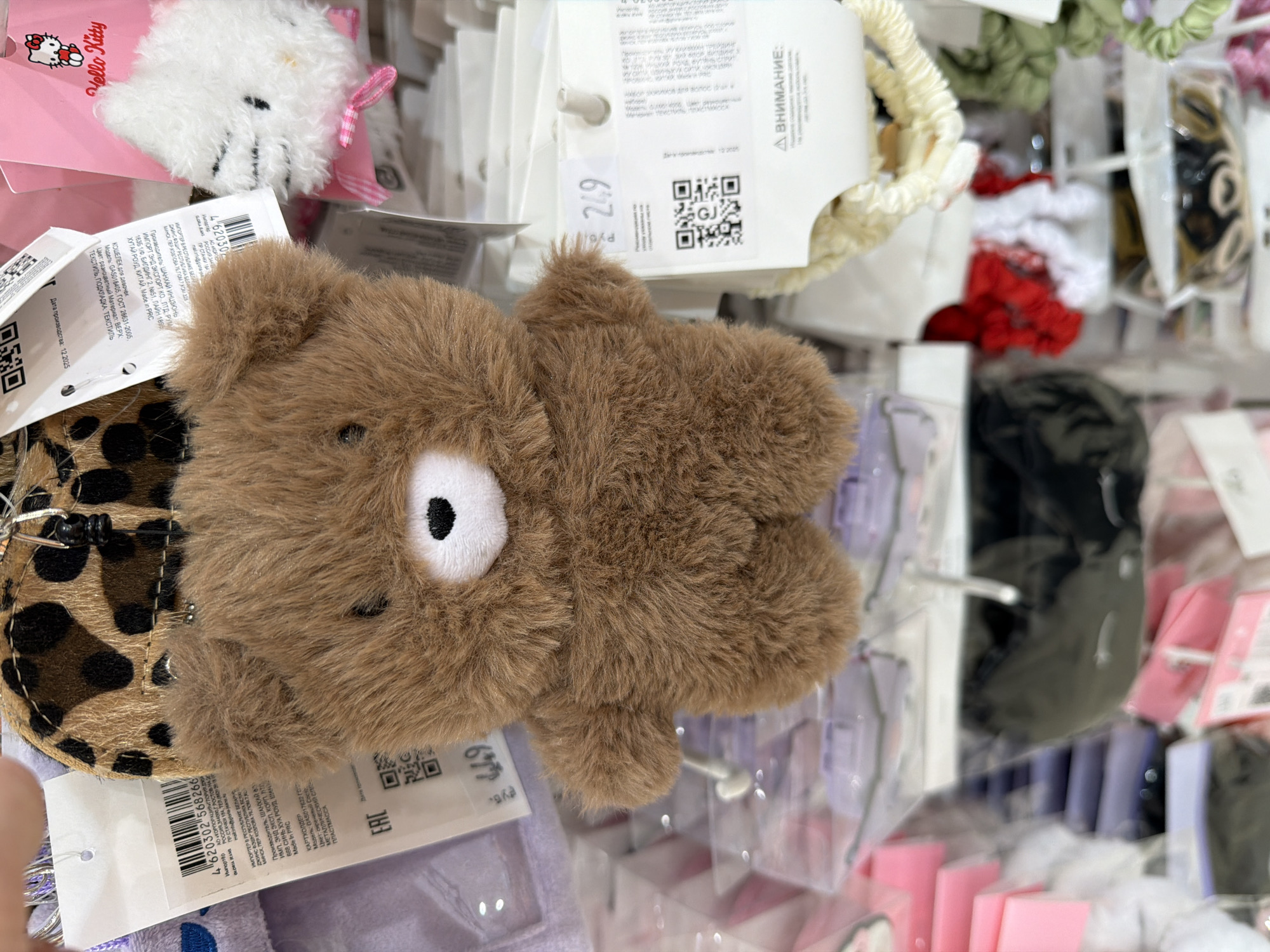 A close-up, slightly angled, indoor shot features a brown teddy bear-shaped coin purse/wallet, seemingly displayed for sale amongst various other small accessories. The bear has a white snout and small black eyes and is attached to a leopard print fabric. Underneath the coin purse is a product tag with barcode and information in Russian, including model number GASD17622 and GAS018405 and manufacturer information of Shanghai Incjun Import End Export. A Hello Kitty product is also visible, as are a few hair ties. The photograph was likely taken in a retail store or market. The tag indicates that the image location is Berezniki, Russia.
