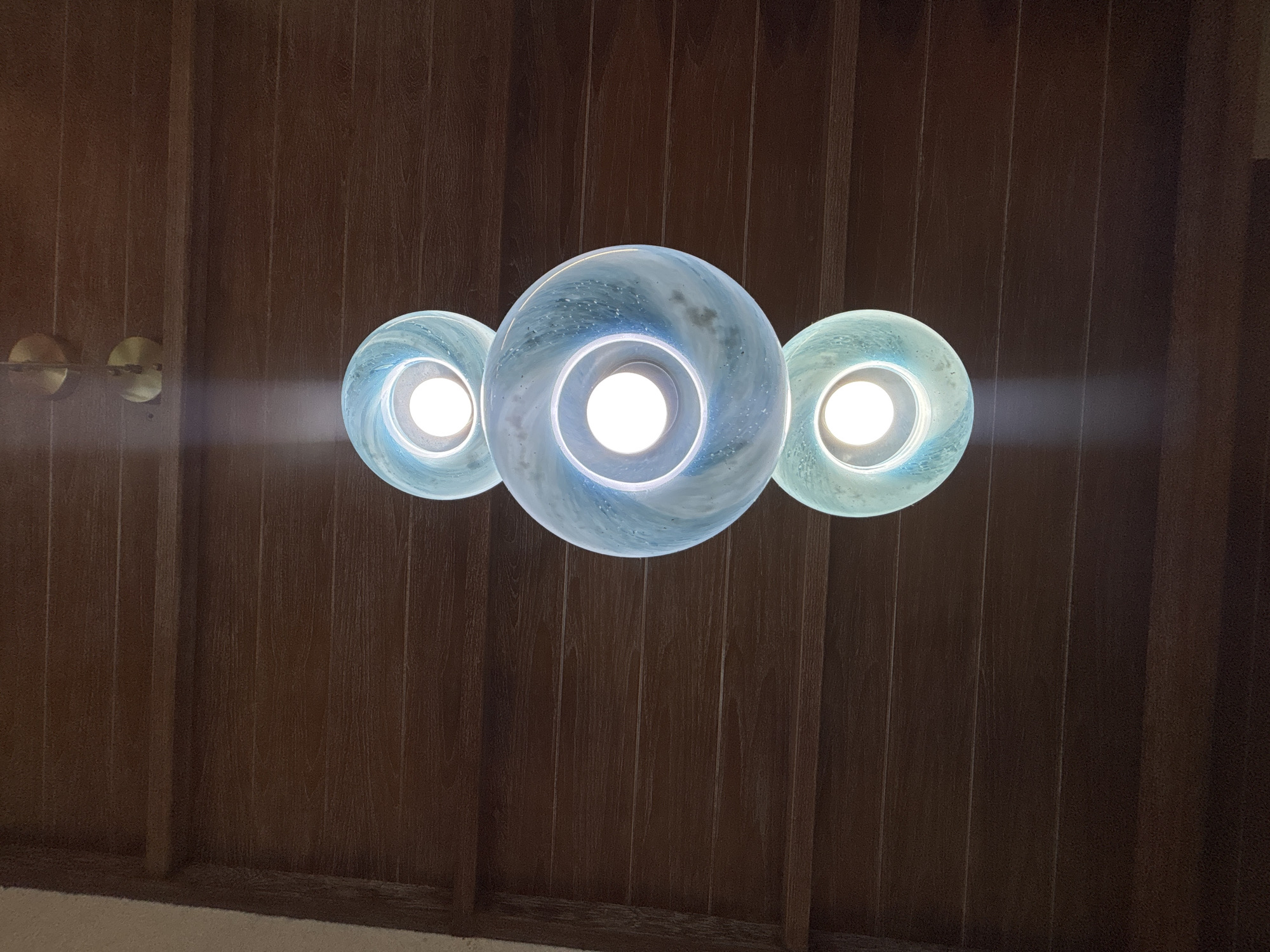 Three spherical, blue-swirled glass pendant lights hang from a dark wood paneled ceiling. The lights are illuminated from within, casting a bright white glow through the translucent glass. Two circular, brass fixtures are visible on the left side of the image, mounted on the ceiling paneling. The scene is indoors, likely a residential or commercial interior in Chalong, Thailand.