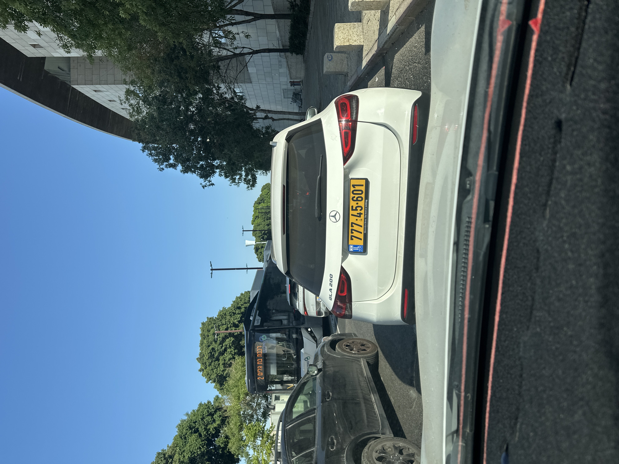 In Haifa, Israel, on a clear day, traffic is stopped on a multi-lane road. A black SUV is on the left, and a white Mercedes-Benz GLA 200 is in the center lane, bearing the license plate 777-45-601. A dark bus with the route number "2" and Hebrew text indicating "Keren Ha'Yam" is in the lane beyond the Mercedes. A white building with a long, angled roof is visible on the right, with trees and a sidewalk. The perspective suggests the image was taken from inside another vehicle.