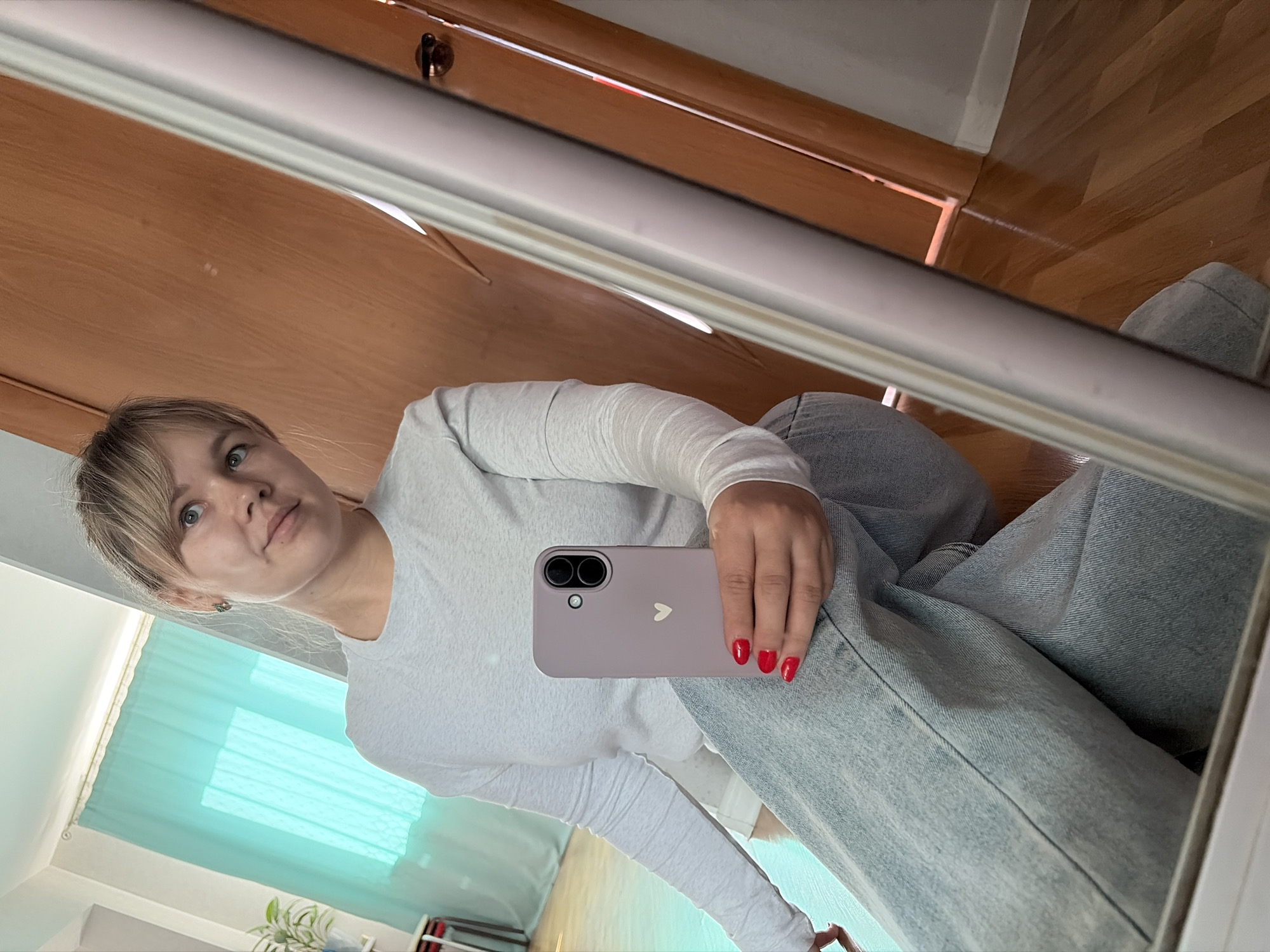 A young woman is taking a selfie in a mirror. She is wearing a long-sleeved white shirt and light blue denim jeans. She is holding a light purple smartphone with a white heart on the back. Her nails are painted red. The background includes a wooden door, a light blue window covering, and a potted plant. Based on the available information, it is not possible to confirm the location in the city of Chaikovsky, Russia.