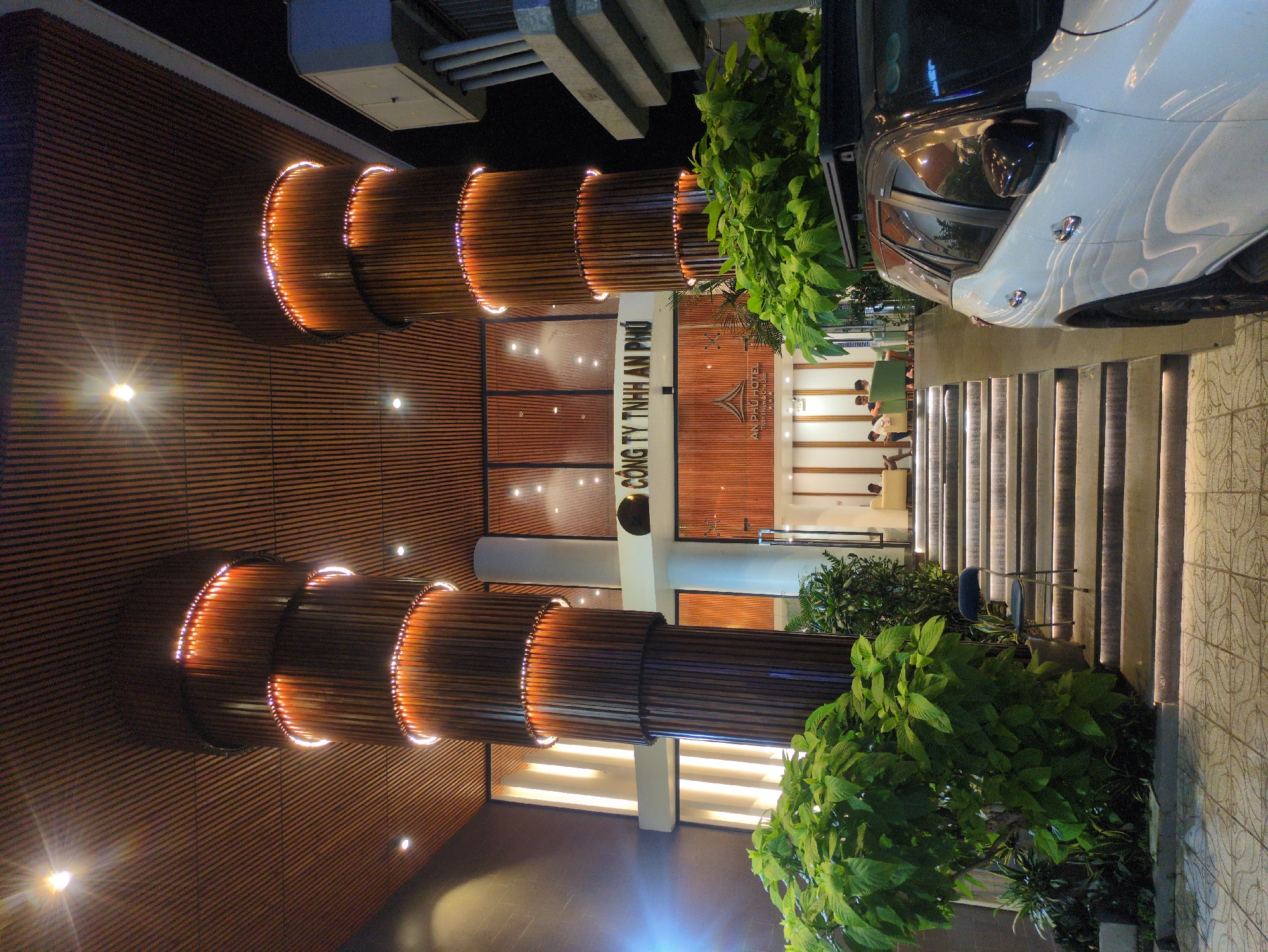 The image shows the exterior of the An Phu Hotel in Phú Quốc, Vietnam. The facade features a horizontal wooden panel design with decorative illuminated cylindrical elements. To the right is a white car parked near a set of illuminated steps. Lush greenery is planted near the stairs. Inside the hotel lobby, a few people are seated. The hotel's name is displayed on the front.