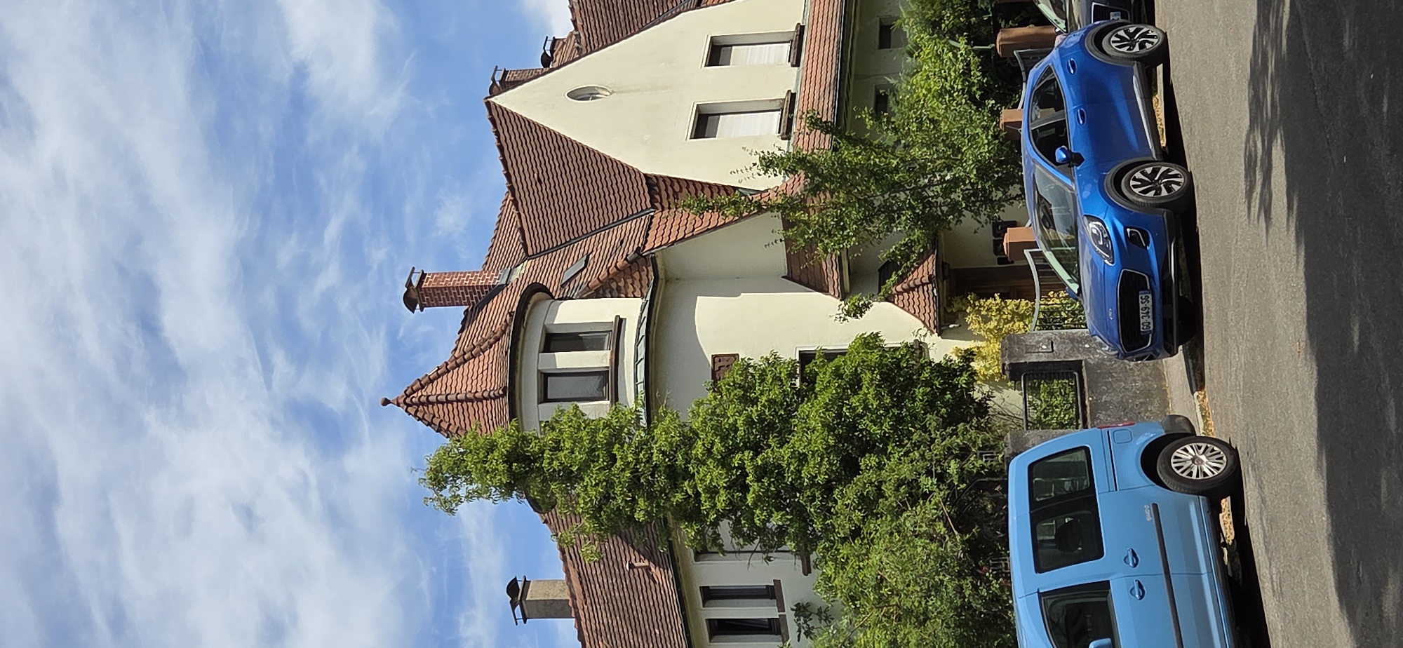 A bright blue Ford Puma is parked on a residential street in Kehl, Germany, next to a bright blue van. The cars are parked in front of a large, two-story house with a red-tiled roof and cream-colored walls. The house has a prominent round turret with a pointed roof. Mature trees and greenery surround the house. The sky is blue with scattered white clouds.