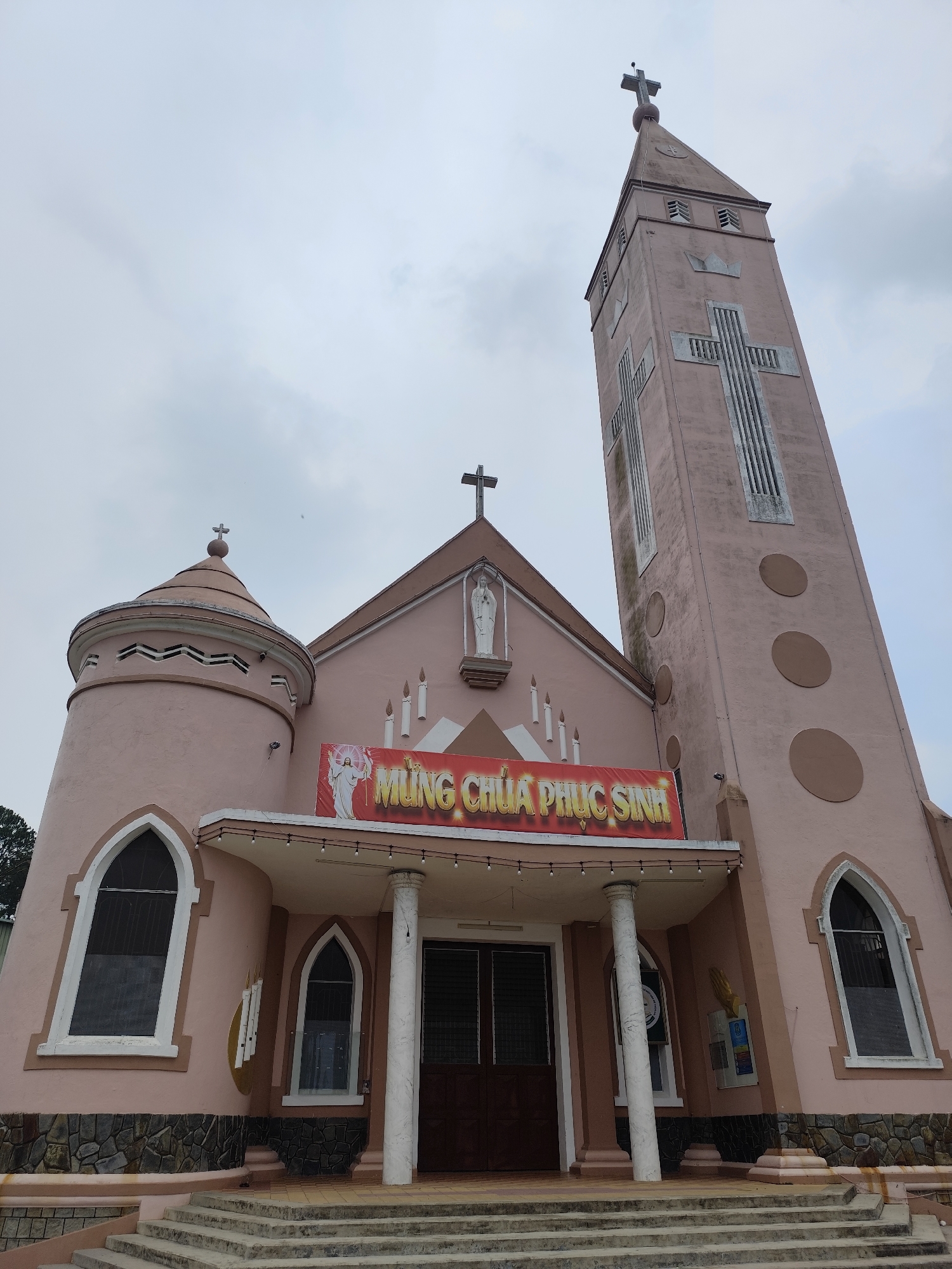 The image shows a church in Lang Biang - Đà Lạt, Vietnam. The building is painted a light pink color and has a tall tower with a cross on top. A large banner with the words "MUNG CHÚA PHỤC SINH" is hung above the entrance. The church has a set of stairs leading up to the main doors, and several windows are visible on the sides of the building. The sky is overcast and gray. The architecture is a mix of European and Vietnamese styles.