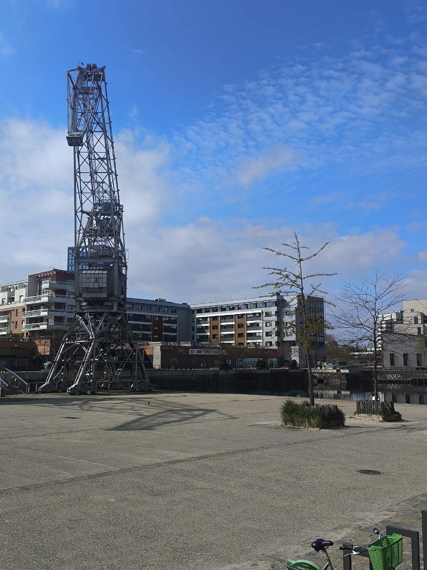 The image depicts a sunny, daytime scene in Strasbourg, France, featuring the "Grue Jaune" (Yellow Crane) or "Grue à Tour" (Tower Crane), a historical port crane. The crane, constructed of steel, stands tall against a bright blue sky with scattered clouds. Modern apartment buildings are visible behind the crane. In the foreground is a large paved area with a few trees and some low plantings surrounded by fencing. A canal or waterway is visible behind the trees. A green bicycle is visible on the bottom-right corner of the image. No people are visible in the frame.