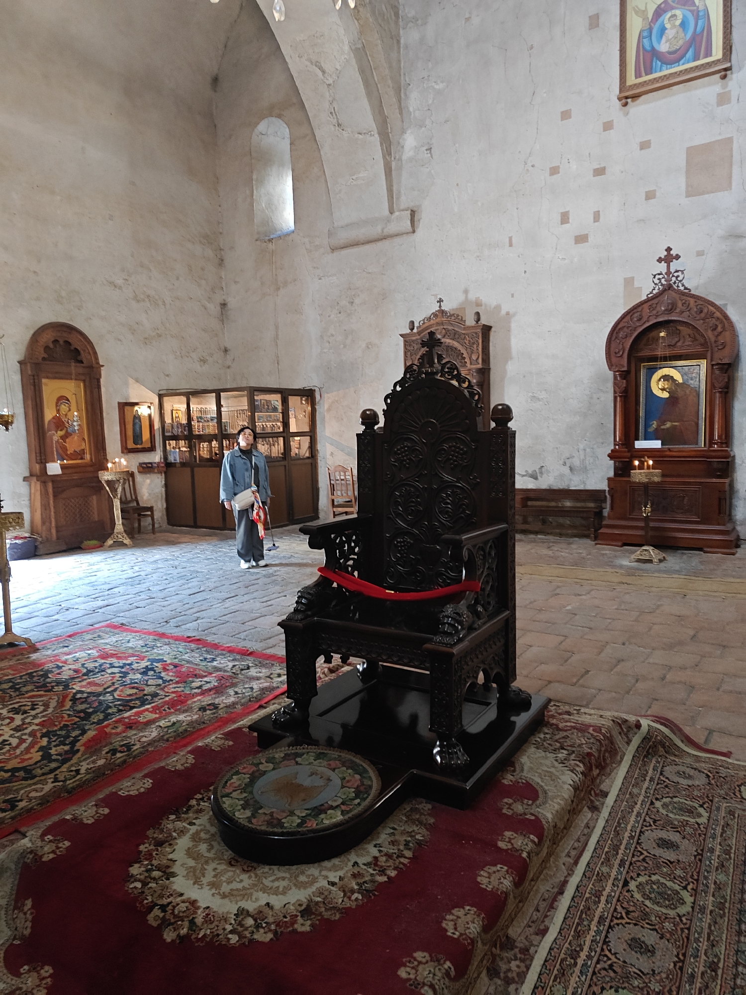 A full-length shot shows the interior of Svetitskhoveli Cathedral in Pkhundavi, Georgia. A person, presumably female, dressed in a light blue jacket, grey pants, and white sneakers, with a white bag slung over her shoulder, stands towards the left of the frame. In the foreground, a large, ornate, dark wood chair with a red ribbon is set upon a patterned rug. The cathedral interior features exposed brick flooring and rough white walls. Religious icons are displayed in wooden frames on the walls. A window is visible in the background above an arch.