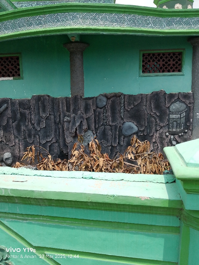 The image shows a close-up view of a section of a building in Kecamatan Haurgeulis, Indonesia. The building is painted a bright green and has a stone-like patterned facade. The facade is made up of small grey stones and features an intricate design of a mosque. Above the facade, a green decorative band lines the edge of the building. The building has a window on the right side of the image that has a decorative design and is also painted a vibrant green. There are dried plants visible beneath the facade. The image was taken at 12:44 PM on March 23, 2023.