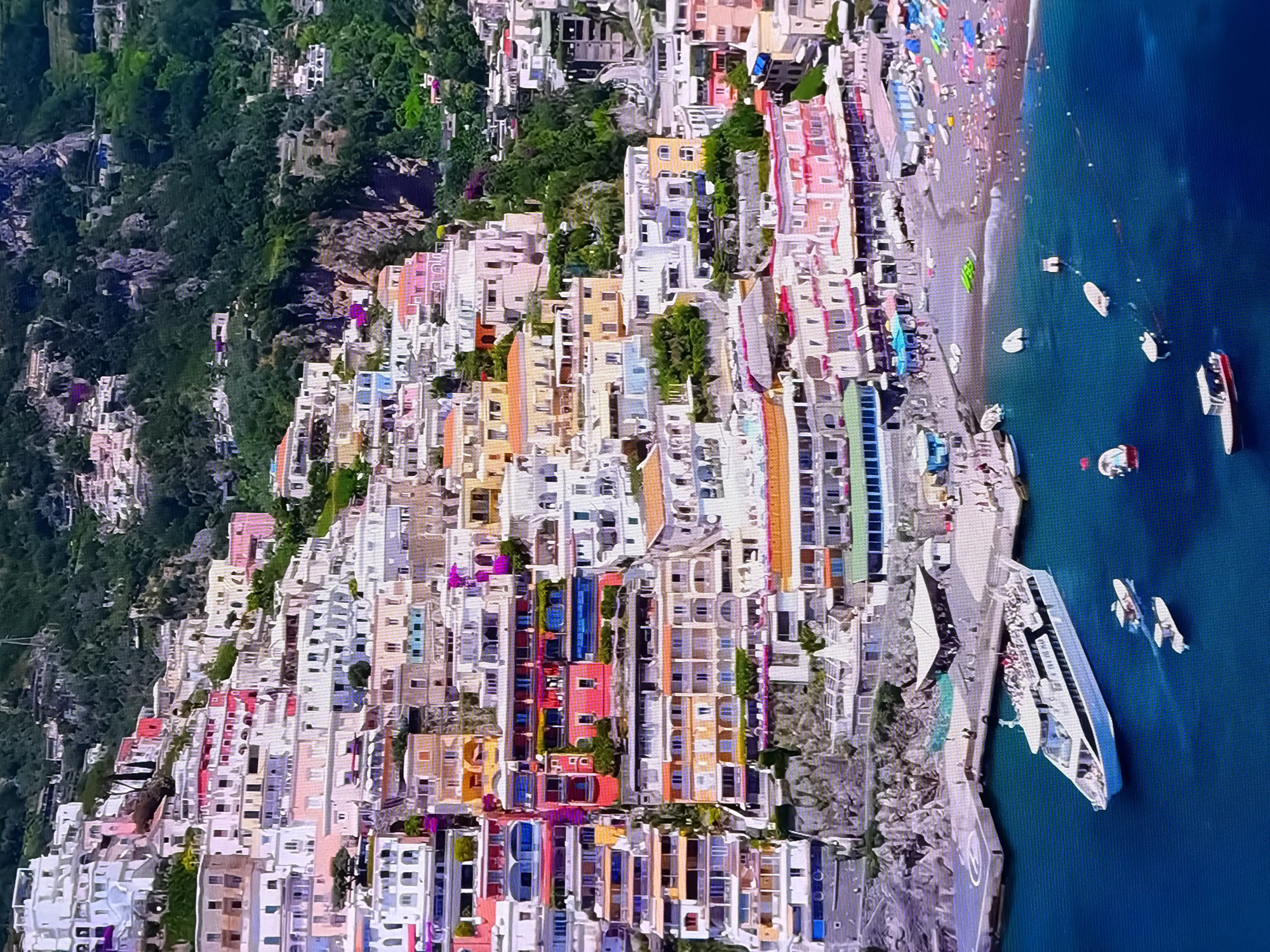 This is an aerial, high-angle shot of a densely packed, colorful town built on a steep hillside overlooking a bright blue body of water. The buildings are a mix of pastel colors like pink, yellow, and white, and they appear to be stacked very close together, following the contour of the land. Lush green trees and rocky terrain are visible on the higher parts of the hillside.
Along the waterfront, there is a beach with many people sunbathing and sitting on lounge chairs. Several boats are visible in the water: a large white ferry-like vessel is docked at a pier, and smaller boats are scattered throughout the bay. The water appears calm and clear.
The scene suggests a popular tourist destination, likely during a sunny day, given the presence of beachgoers and the bright lighting. The architecture and the vibrant colors of the buildings are a prominent feature. There is no discernible text visible in the image.