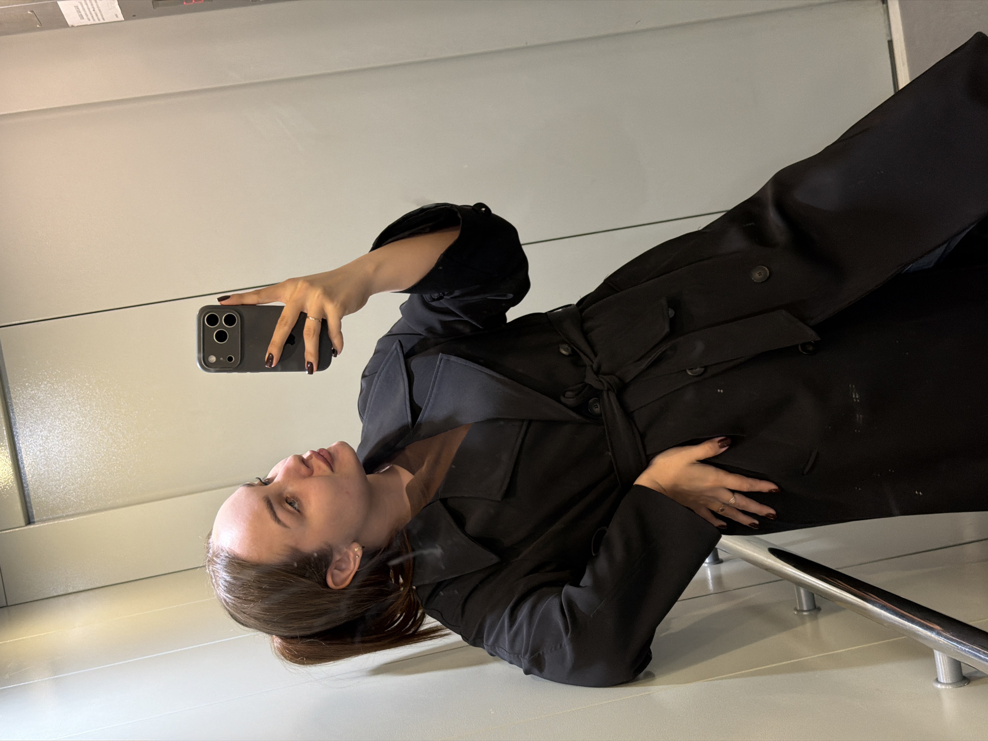 A young woman is seen taking a selfie in what appears to be an elevator. She is wearing a black trench coat and holding a dark-colored smartphone in her right hand. The elevator's interior is simple, with a light-colored floor and walls. A metal handrail is visible in the foreground. There are no visible indicators to suggest the location is Chaikovsky, Russia.