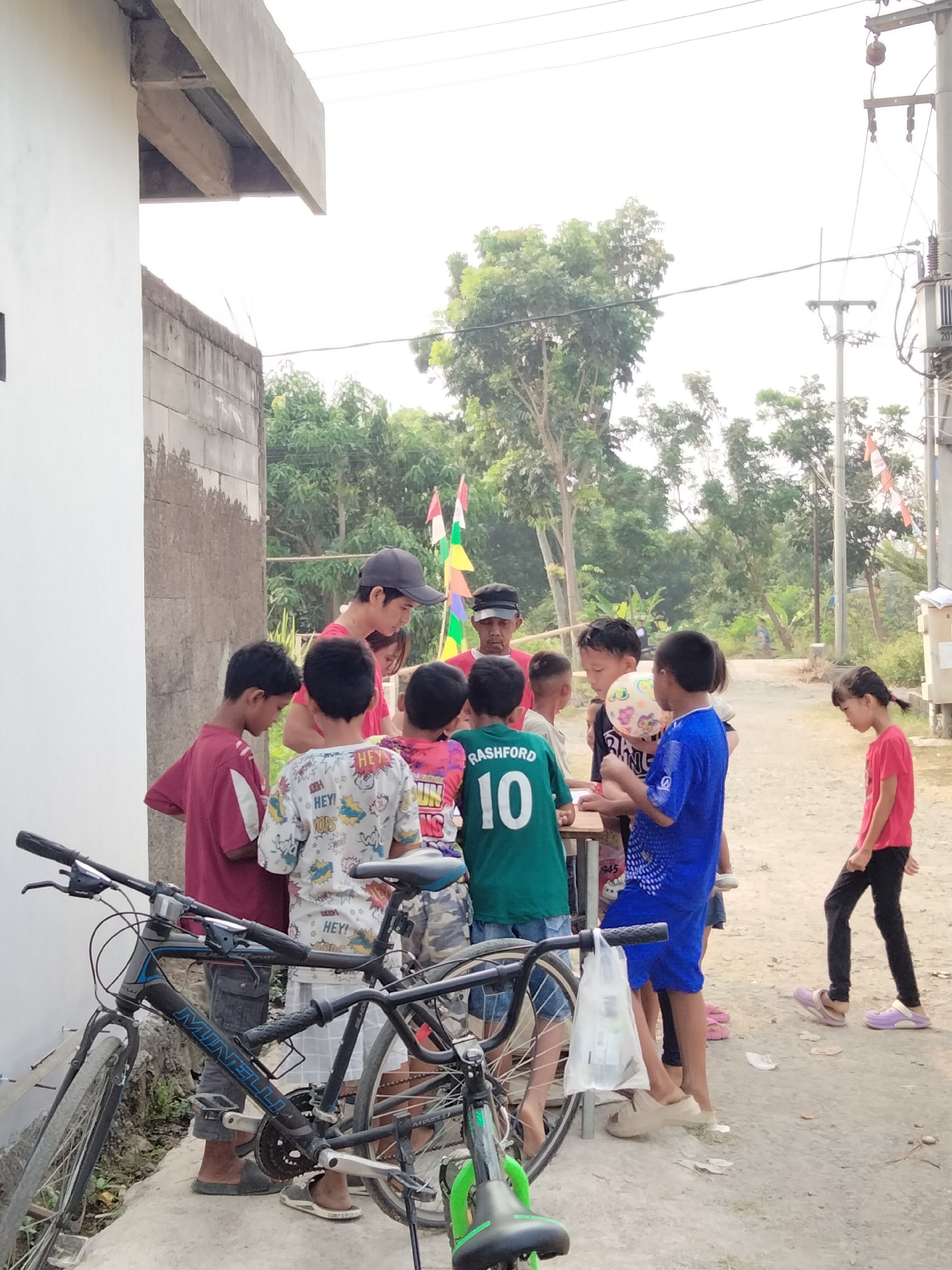 In Cariu, Indonesia, a group of approximately ten children and two adults are gathered outdoors. The children appear to be of various ages, with most wearing casual clothing. One boy in the center is holding a colorful ball. Several children are looking down at something on a low table or surface, possibly interacting with treats or activities. There are two bicycles parked in the foreground. The background features trees, power lines, and utility poles, suggesting a semi-rural or residential area. The scene is illuminated by natural daylight.