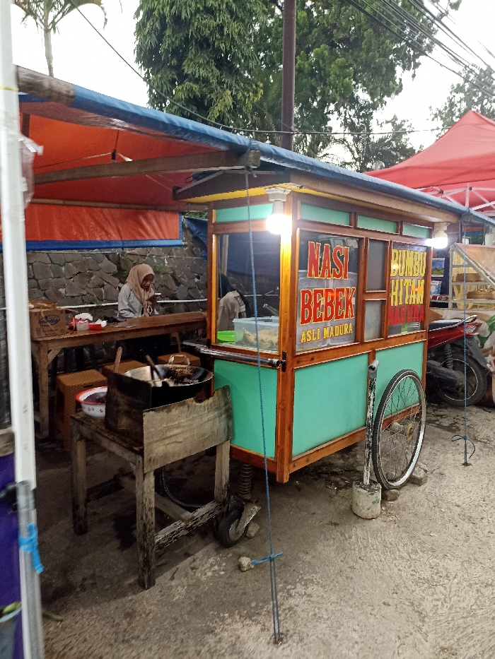 The image shows a street food stall located at the coordinates -6.935 latitude, 107.773 longitude in Jatinangor, Indonesia. The stall is a wooden cart painted turquoise with a sign on the front that says "Nasi Bebek Asli Madura," which translates to "Duck Rice from Madura." The stall is surrounded by other buildings, including a building with a red awning. A woman wearing a white headscarf is sitting at a table, which is directly to the left of the food cart. She is looking down at a device in her hands. On the left side of the stall, a small, wooden table with a stove and a large frying pan is located. A motorcycle and scooter are parked nearby. There is an orange awning above the scene. There are a few wooden beams under the awning, which are holding up the structure. The ground is a gray, concrete surface with a small amount of dirt and debris. There is a blue rope tied to the stall and stretched across the sidewalk to the right of the stall.