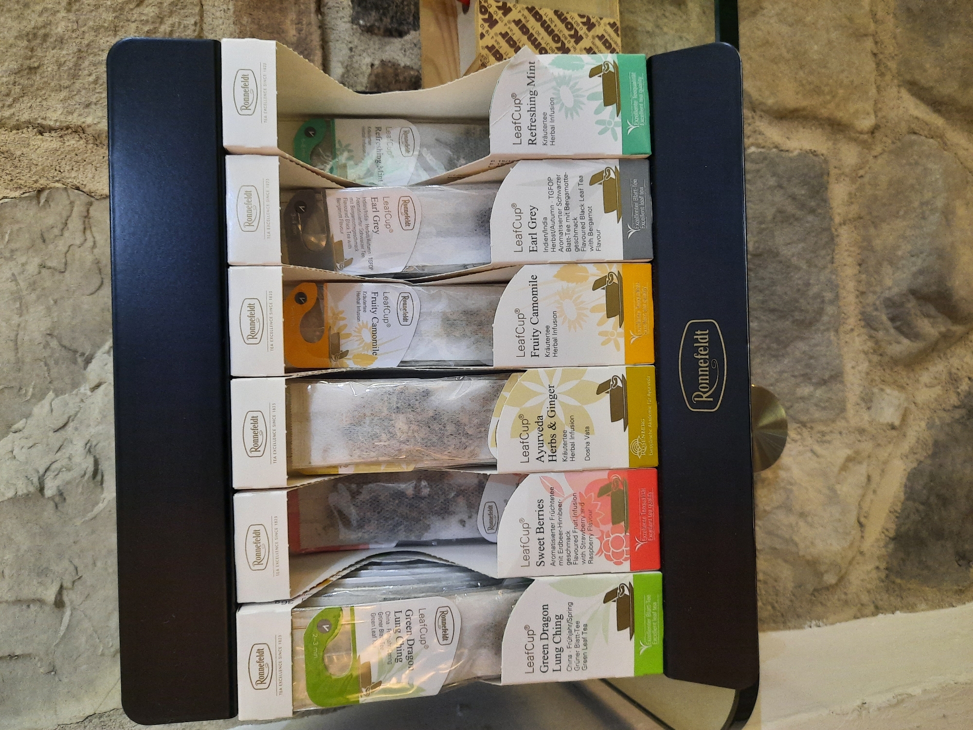 The image shows a display stand for Ronnefeldt LeafCup teas, filled with individual tea sachets. The stand is black and designed to hold multiple rows of tea. The tea boxes are white with colored accents indicating the tea flavor. The background appears to be a stone wall.
Specifically, the visible tea varieties are:
* **LeafCup Green Dragon Lung Ching**: Green tea.
* **LeafCup Sweet Berries**: Flavoured Fruit Infusion.
* **LeafCup Ayurveda Herbs & Ginger**: Herbal Infusion.
* **LeafCup Fruity Camomile**: Herbal Infusion.
* **LeafCup Earl Grey**: Flavoured Black Leaf Tea with Bergamot.
* **LeafCup Refreshing Mint**: Herbal Infusion.
The display is branded with "Ronnefeldt" and "Tea Excellence Since 1823". One of the tea boxes, "Ayurveda Herbs & Ginger", also has "Rosenberg Europäische Akademie für Ayurveda" printed on it, suggesting a connection to an Ayurvedic academy in Rosenberg, which is a town in Bavaria, Germany, but not Rosenheim. The image itself does not provide definitive proof of the location being Rosenheim. The items are arranged neatly, implying a retail or hospitality setting.