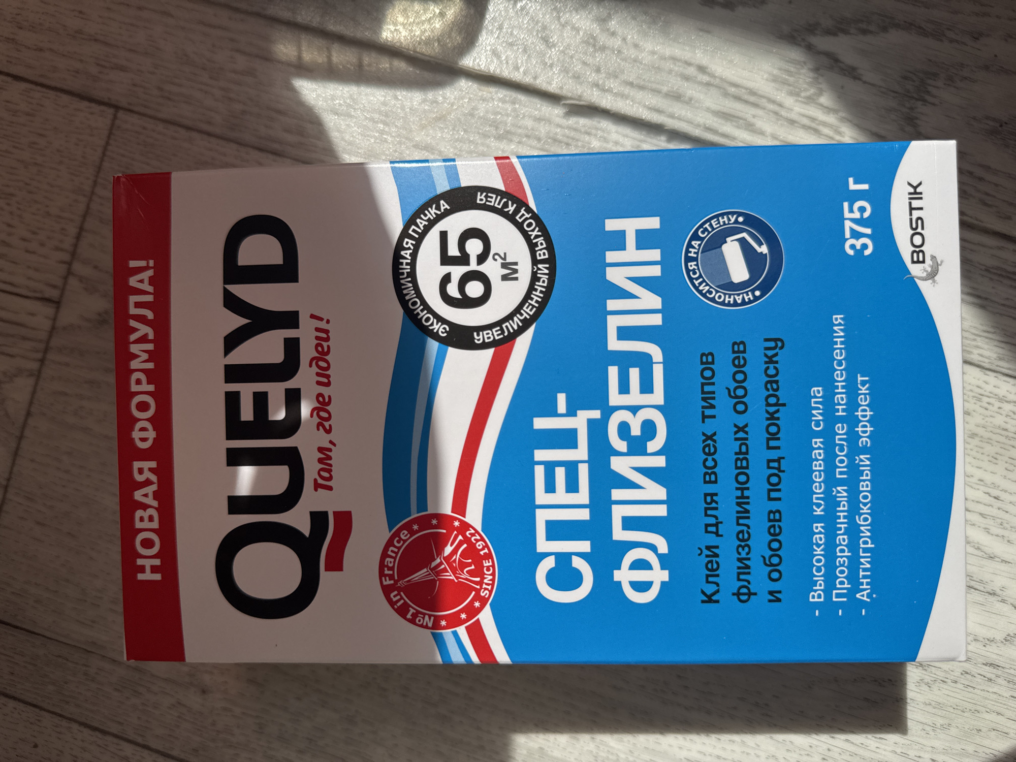 The image shows a close-up of a box of "QUELYD" wallpaper paste. The box is predominantly blue with white and red accents. The branding "QUELYD" is in large black letters at the top. Below it, in smaller red text, it says "Там, где идеи!" which translates to "Where ideas!". There is a circular logo on the left that says "No 1 in France Since 1922" with an image of the Eiffel Tower. On the right, there is another circular logo with Russian text that says "ЭКОНОМИЧНАЯ ПАЧКА" (economical pack) and "УВЕЛИЧЕННЫЙ ВЫХОД КЛЕЯ" (increased glue yield) surrounding the number "65" and "M²". Below the main branding, it says "СПЕЦ-ФЛИЗЕЛИН" (special non-woven wallpaper paste). Further down, in smaller text, it describes the product as "Клей для всех типов флизелиновых обоев и обоев под покраску" (Glue for all types of non-woven wallpapers and paintable wallpapers). Bullet points list key features: "- Высокая клеевая сила" (High adhesive strength), "- Прозрачный после нанесения" (Transparent after application), and "- Антигрибковый эффект" (Anti-fungal effect). The weight is indicated as "375 г". At the bottom right, the brand "BOSTIK" is visible with its gecko logo. The background appears to be a wooden floor. The image is taken indoors, with natural light casting shadows on the box and floor. The location of Яромінскі сельскі Савет, Belarus, is not discernible from the image.