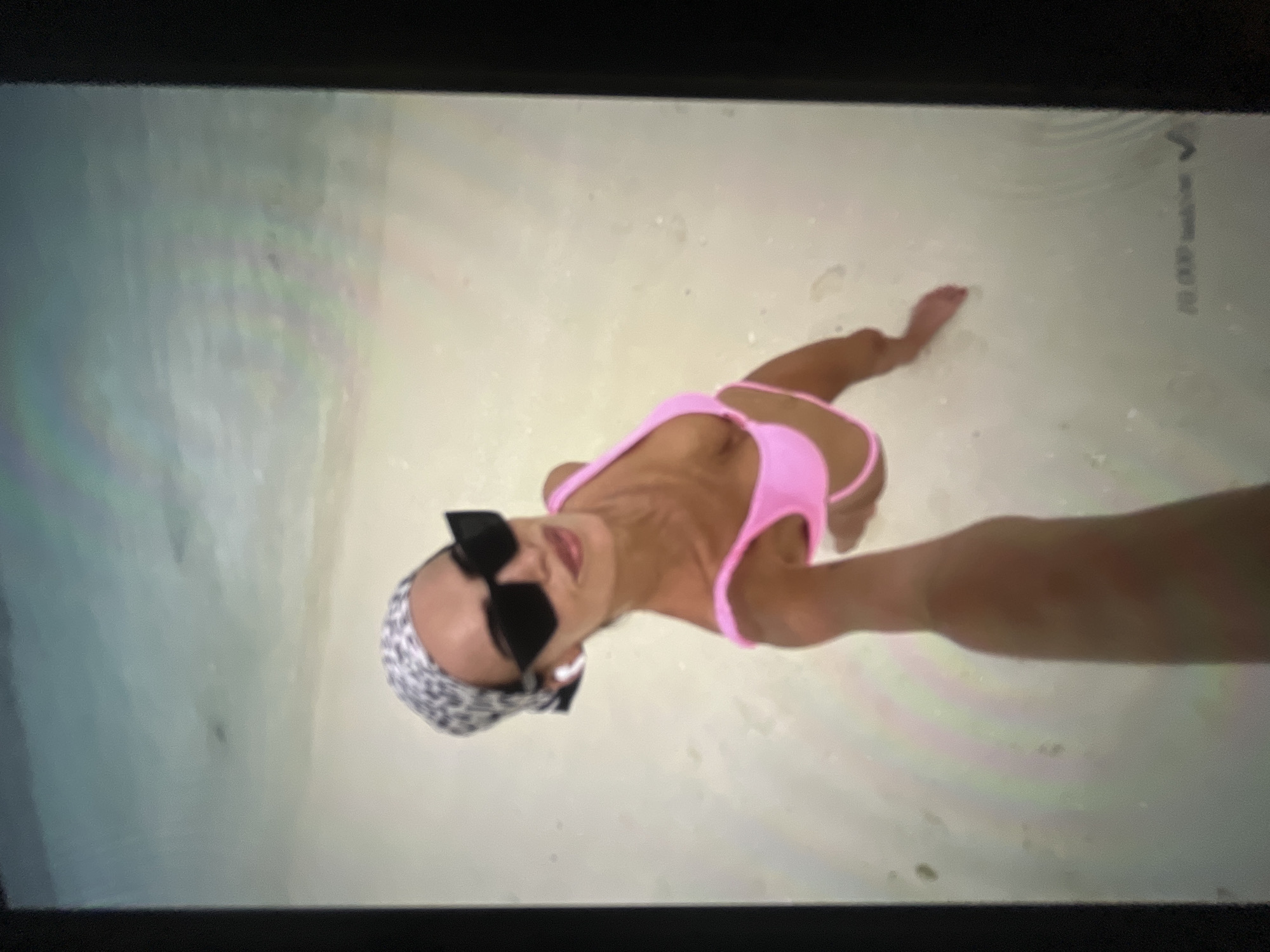 A woman is taking a selfie on a beach in Ravno pole, Bulgaria. She is wearing a pink bikini, black sunglasses, and a leopard-print headscarf. The water is shallow and clear, and the sand is white. The woman is smiling and looking at the camera. The overall scene is bright and sunny.