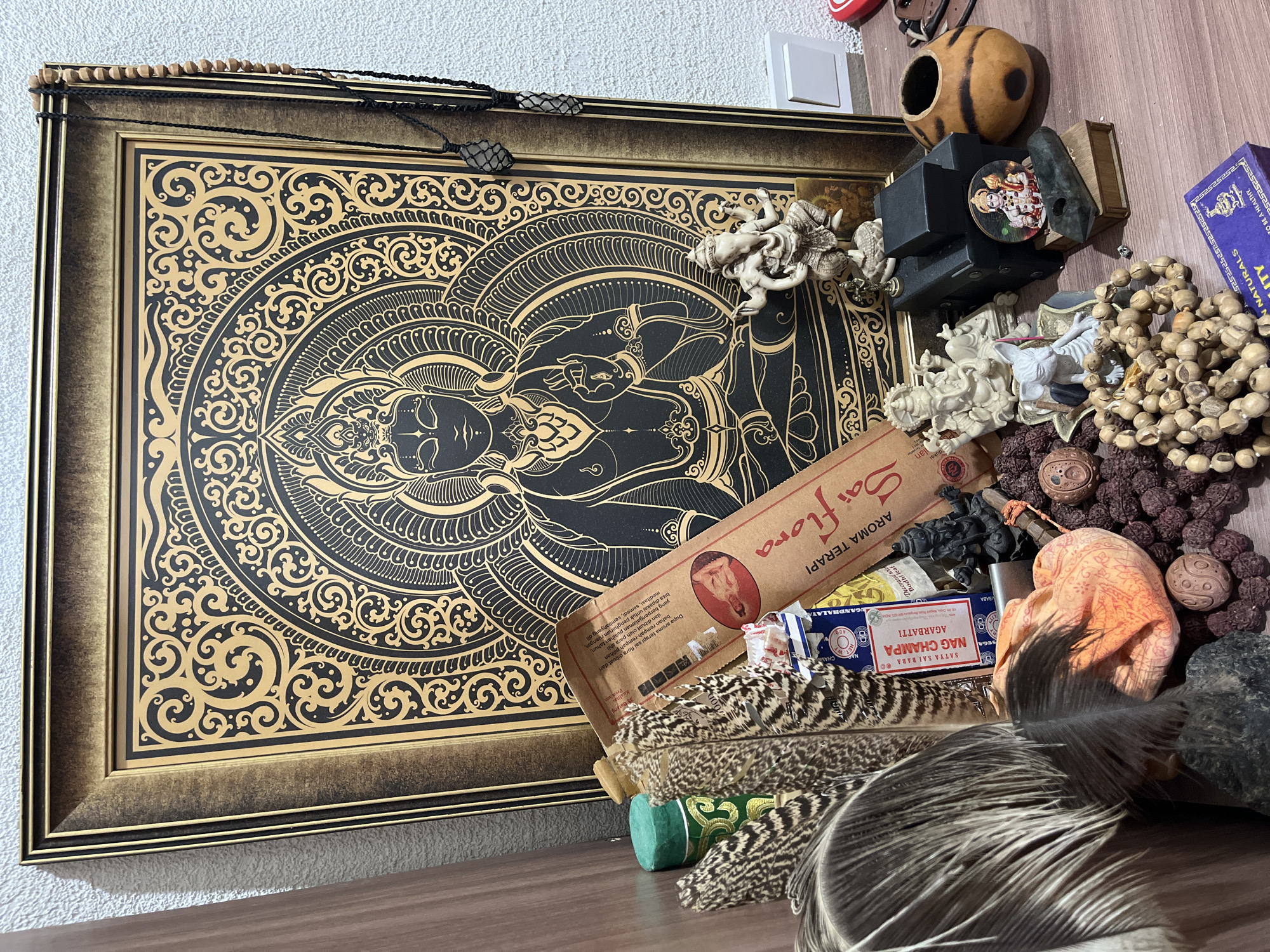 The image shows a shelf with various items related to spirituality and meditation. The central subject is a framed artwork featuring a mandala design with a depiction of what appears to be Buddha. Surrounding the artwork are several objects, including prayer beads, small figurines of deities or spiritual figures, incense sticks, feathers, and a small gourd-like object. The lighting suggests it's daytime indoors, with artificial light casting shadows. The overall mood is one of quiet contemplation and spiritual practice.
Visible text includes: "SATYA SAI BABA", "NAG CHAMPA", "AGARBATTI", "AROMA TERAPI", "Saiflora", and "Bodhi leaf". The location context provided is "Balaklavskyi District, Ukraine", however, there are no visual cues in the image to confirm this specific location. The objects themselves suggest an Eastern or spiritual theme, possibly from India or Southeast Asia. There are no people visible in the image.