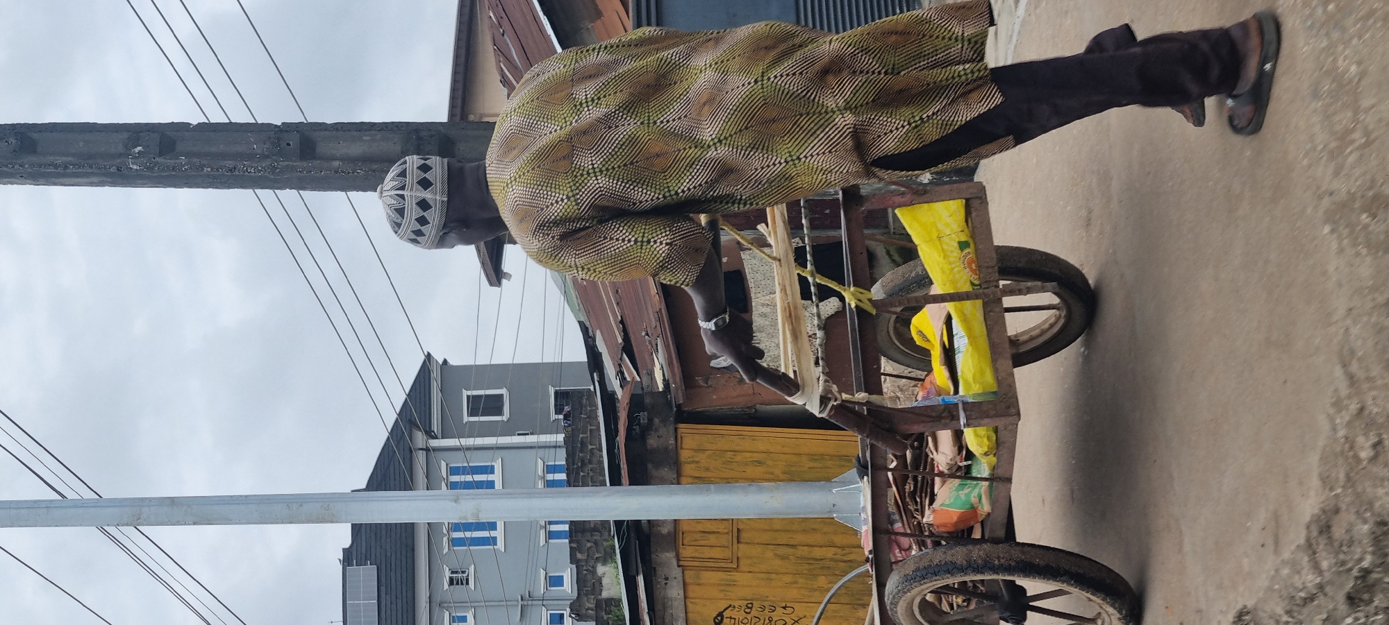 In Ikeja, Nigeria, a man wearing a traditional patterned shirt and a patterned cap pushes a two-wheeled cart loaded with various items, including yellow plastic bags and what appear to be bundles of papers or cardboard. The cart has a metal frame and wooden components. The man is seen from behind, and his right hand is on the cart's handle. He is wearing dark pants and sandals. In the background, a grey multi-story building with blue-tinted windows stands behind a collection of weathered, corrugated metal roofs. A tall, grey utility pole with electrical wires running across it is prominent in the mid-ground, near the man and the cart. The scene is set on a paved surface, likely a street, under an overcast sky.
